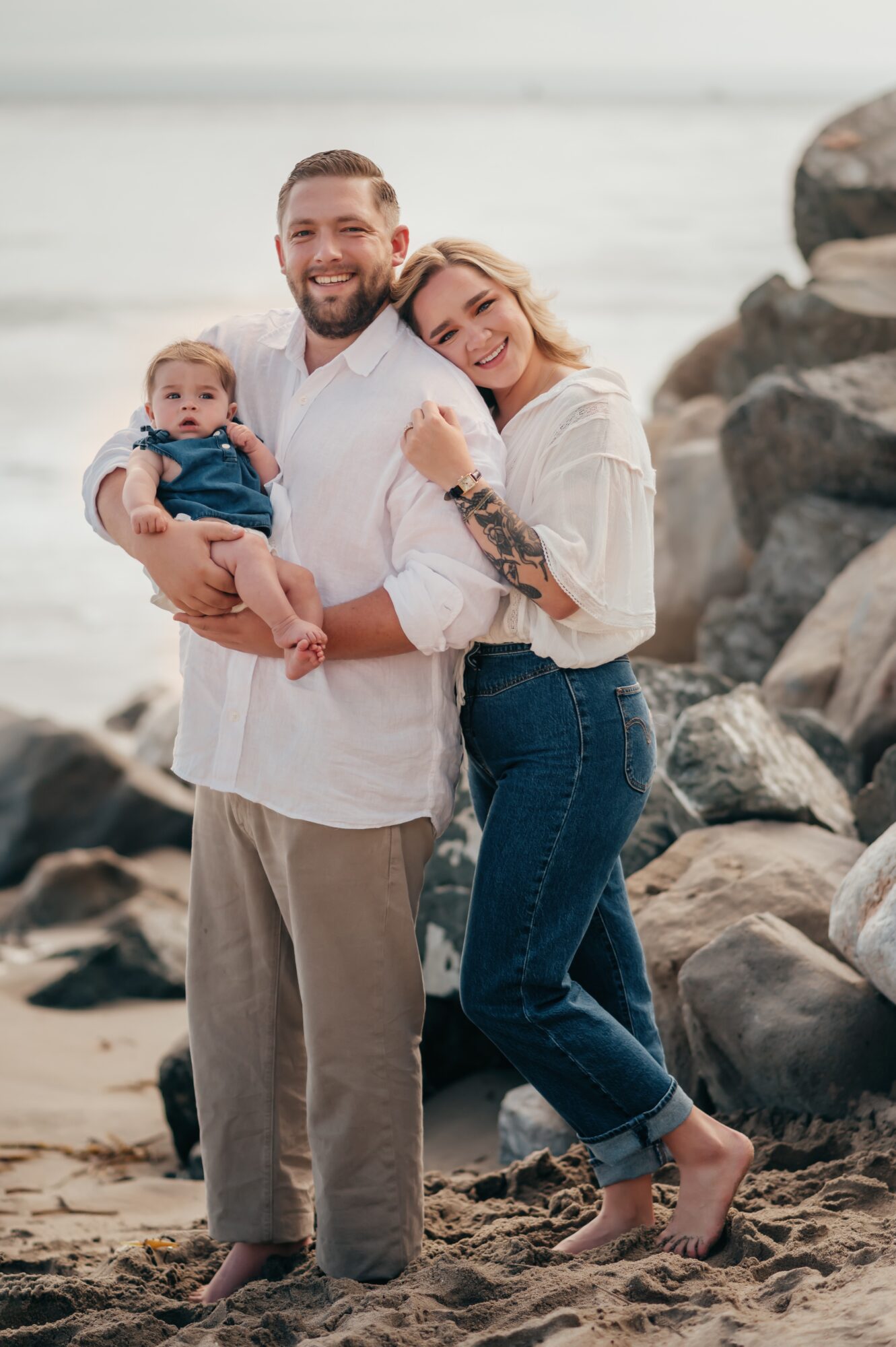 Family of three on a rocky beach, smiling, with a man holding a baby and a woman leaning on him, ocean in background.
