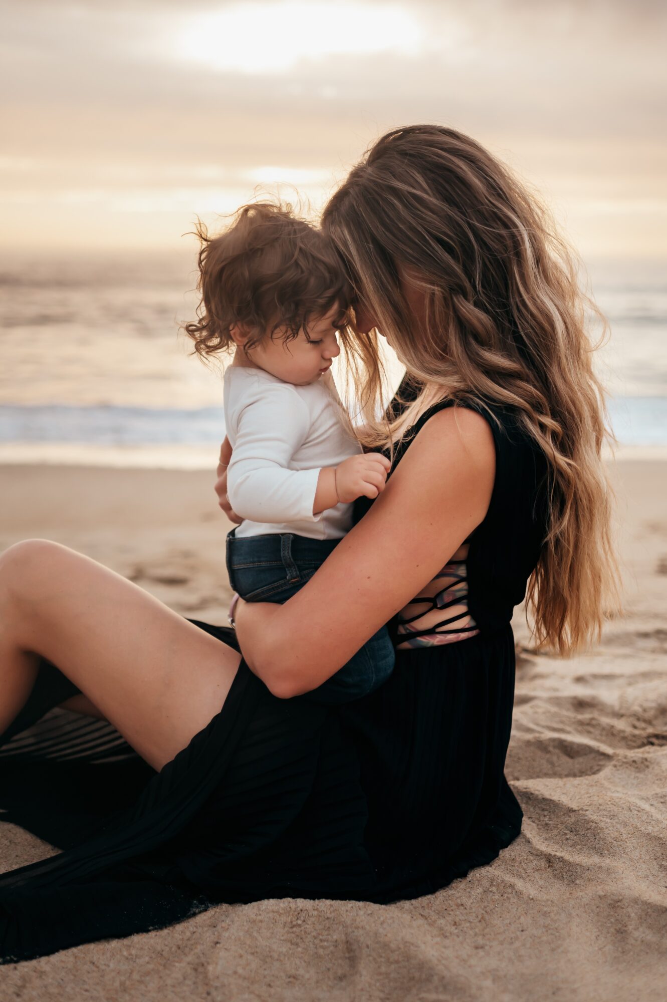 Woman sitting on sand holding a child, both with heads close together, at the beach during sunset