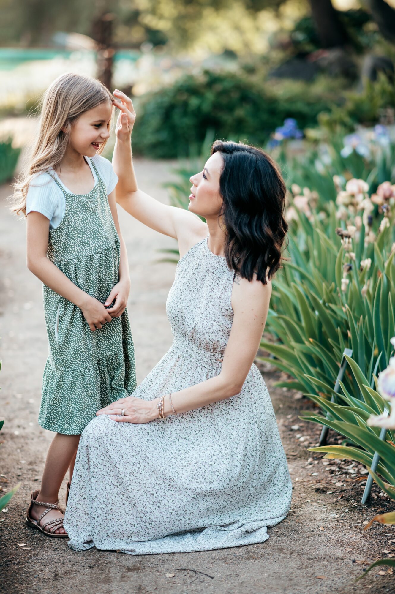 Woman kneeling and touching a young girl's forehead in a garden with flowers and greenery.