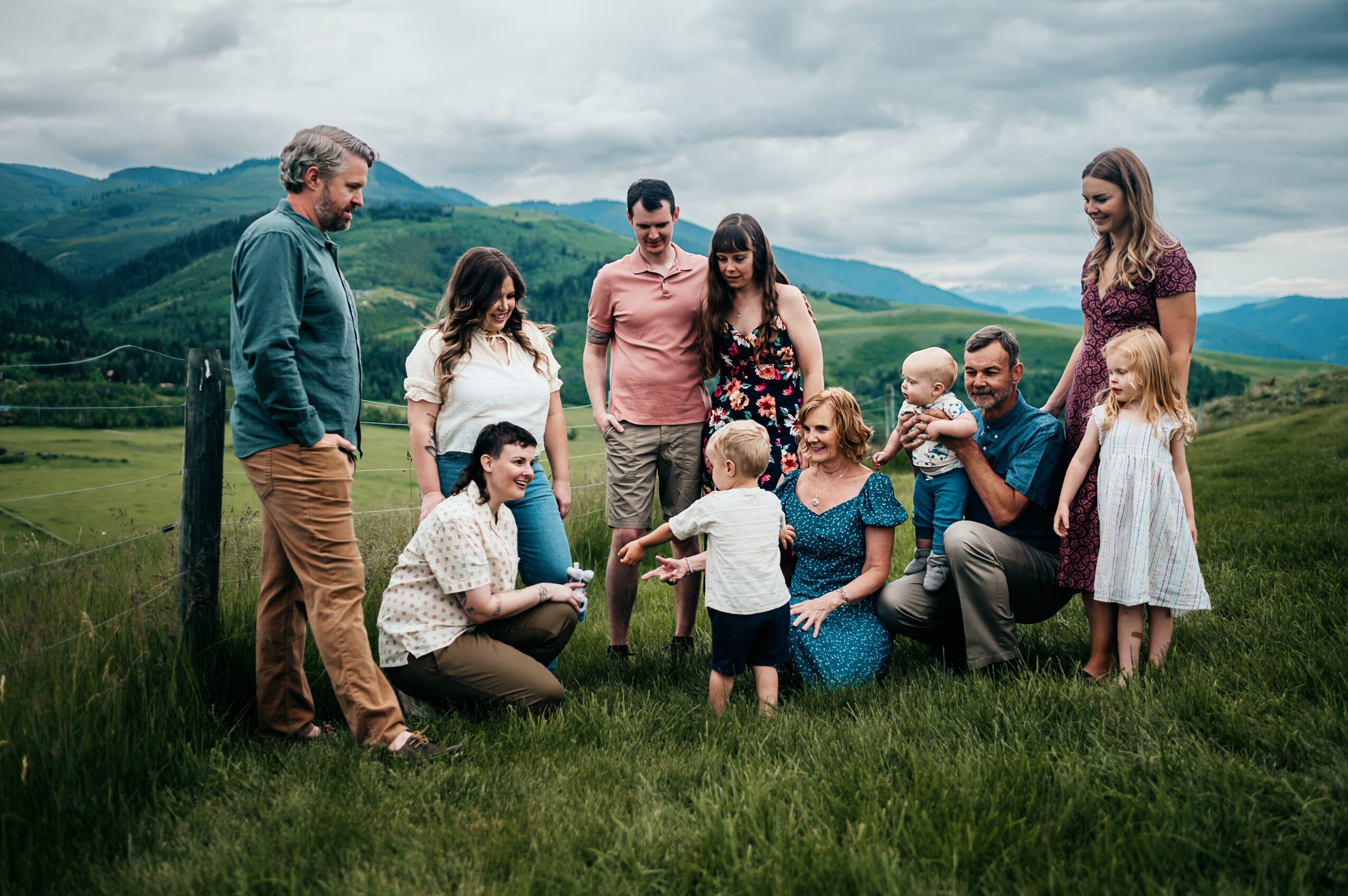 Group of people outdoors in a grassy field with mountains in the background, some standing and some sitting, children present.