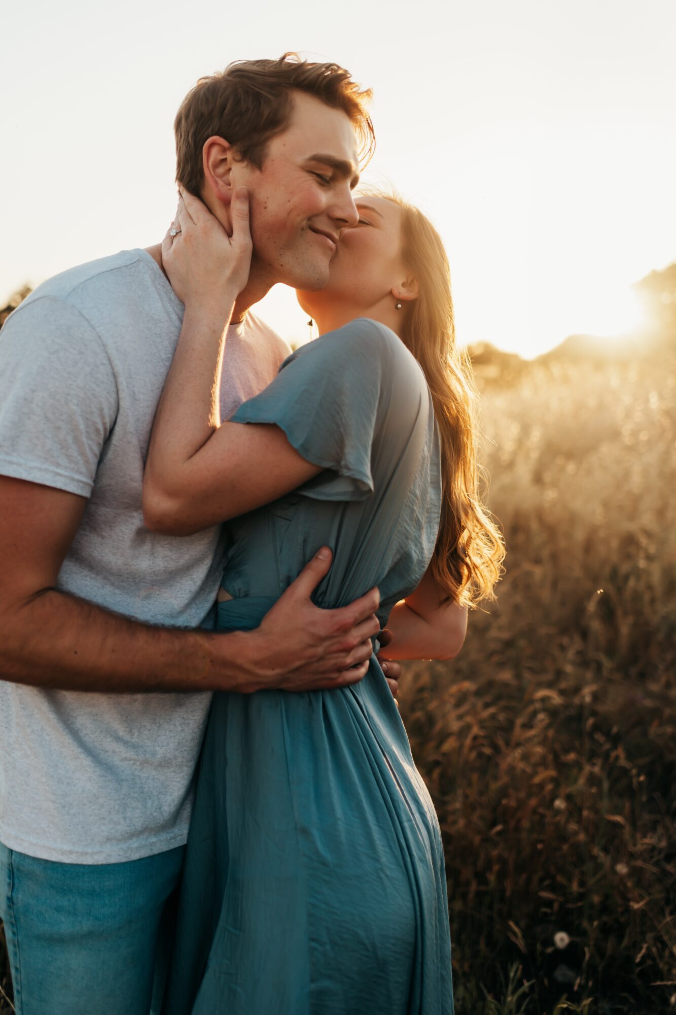A couple embraces outdoors during sunset, with the woman kissing the man's cheek, both smiling, in a field.