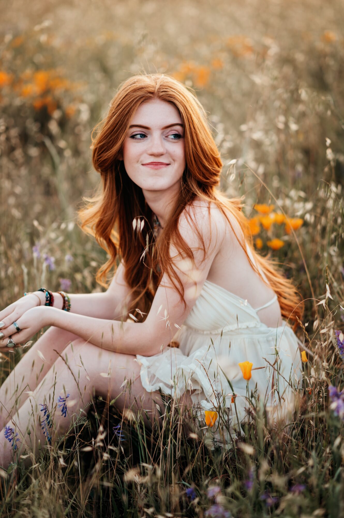 Young woman with long red hair sitting in a field of wildflowers, smiling softly, wearing a white dress.