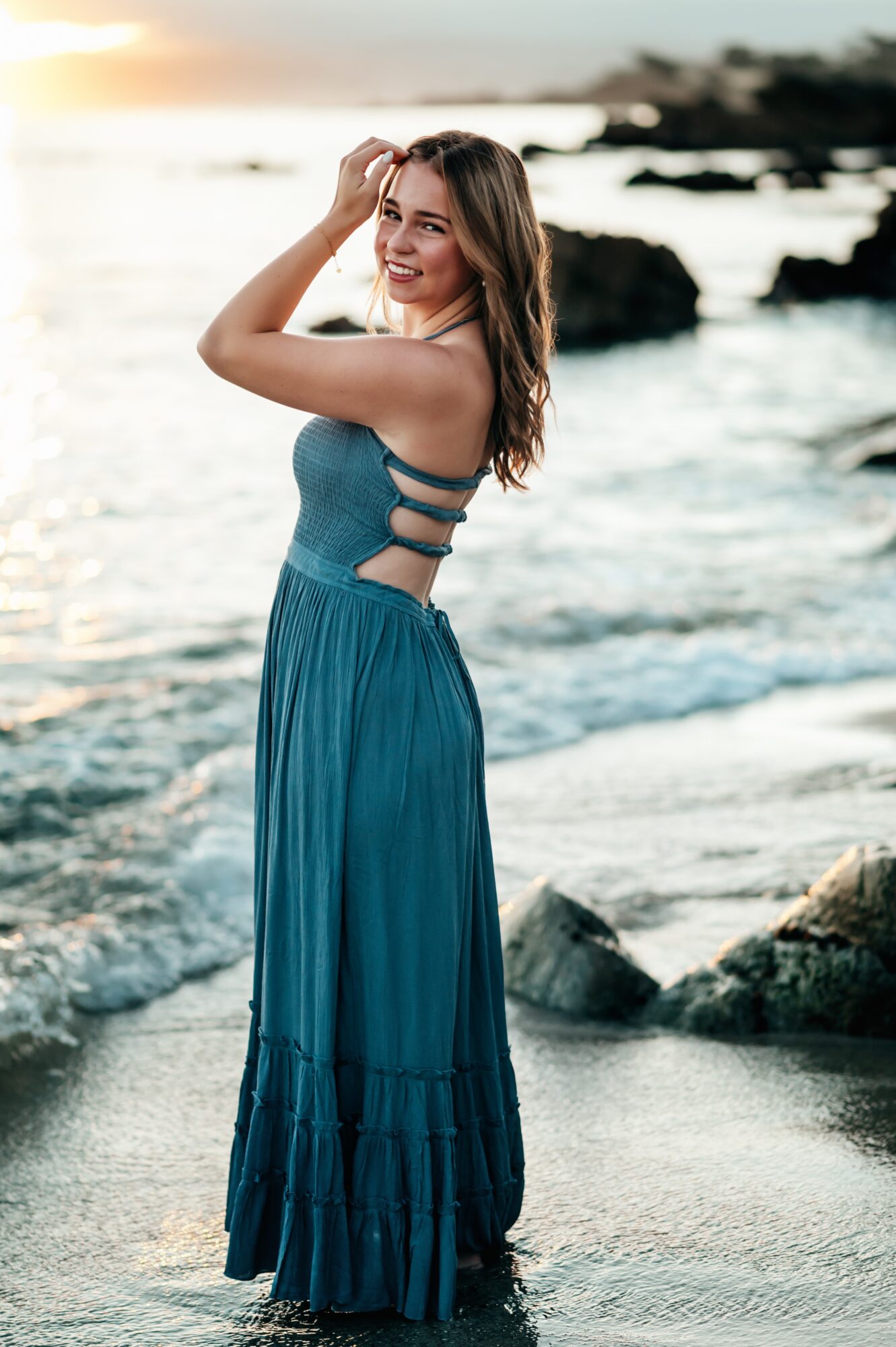 Woman in a blue dress standing on a beach with rocks and ocean waves, smiling, during sunset.