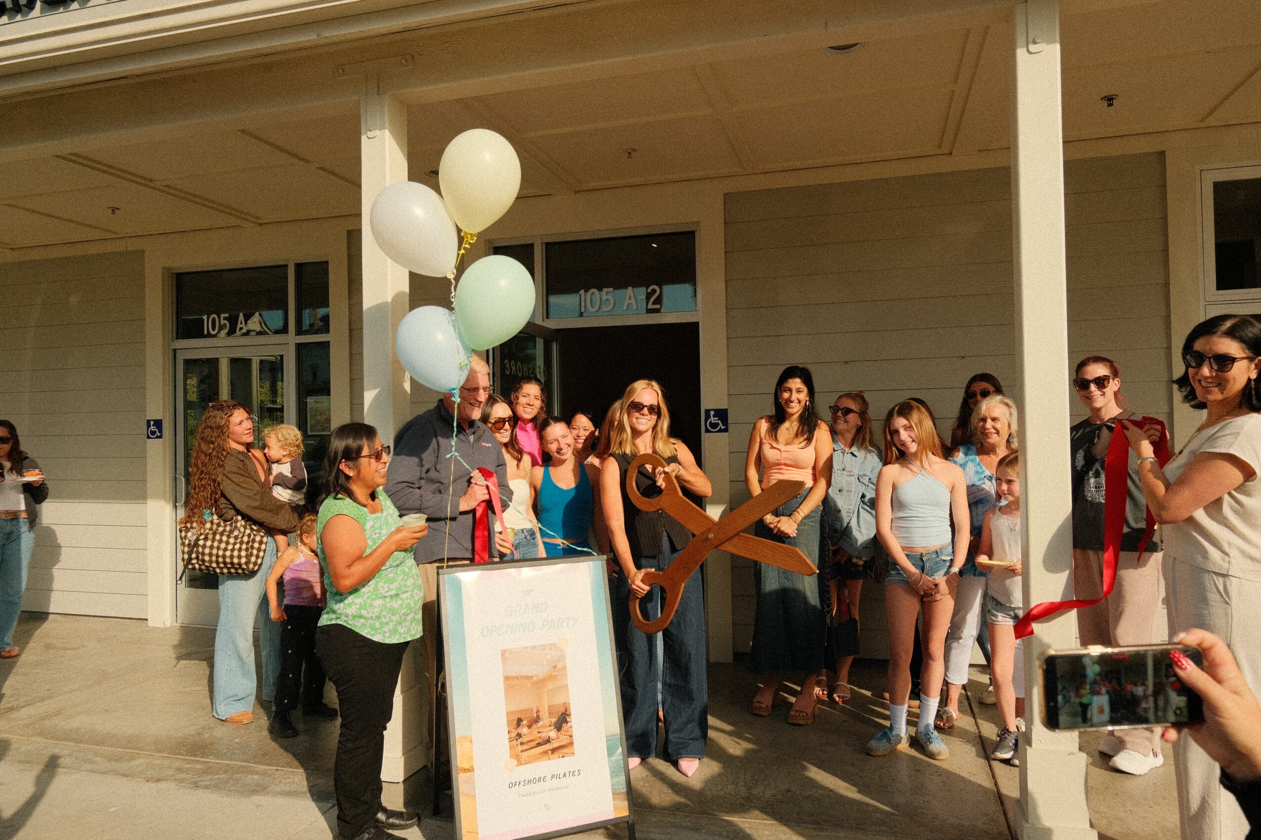 Group of people gathered outside a building for a ribbon-cutting ceremony, with balloons and a large pair of scissors.
