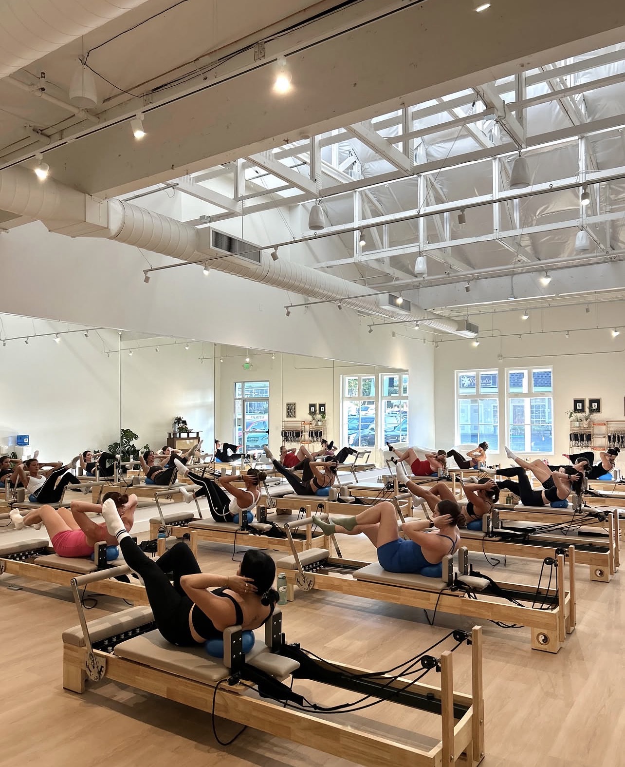 People exercising on Pilates reformer machines in a bright, spacious gym with large windows and high ceilings.