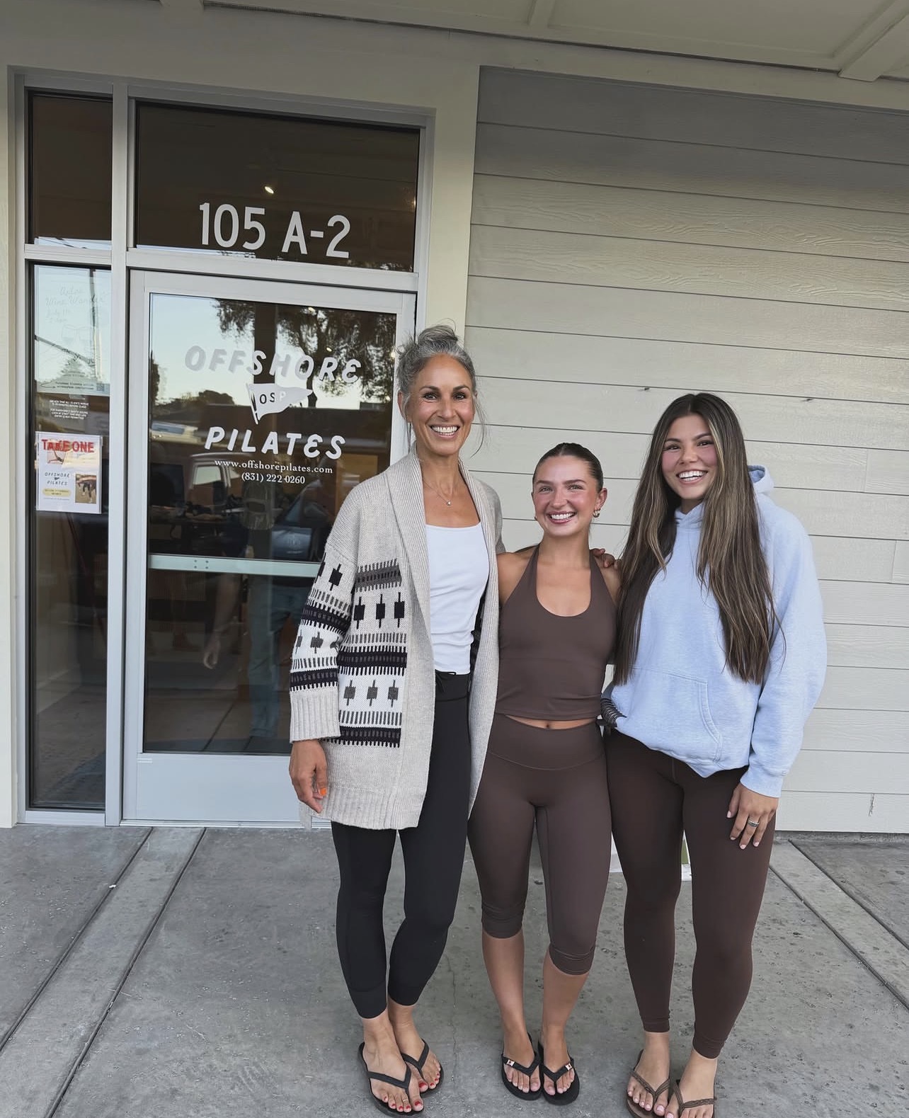 Three women standing outside a building with a glass door, smiling, with signs on the door, one wearing a patterned cardigan.
