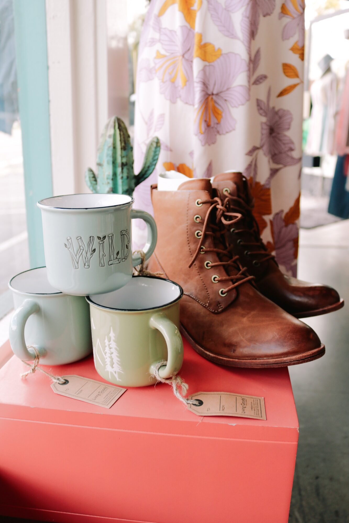 Brown leather boots, two ceramic mugs, and a cactus plant on a pink surface, with floral curtains in the background.