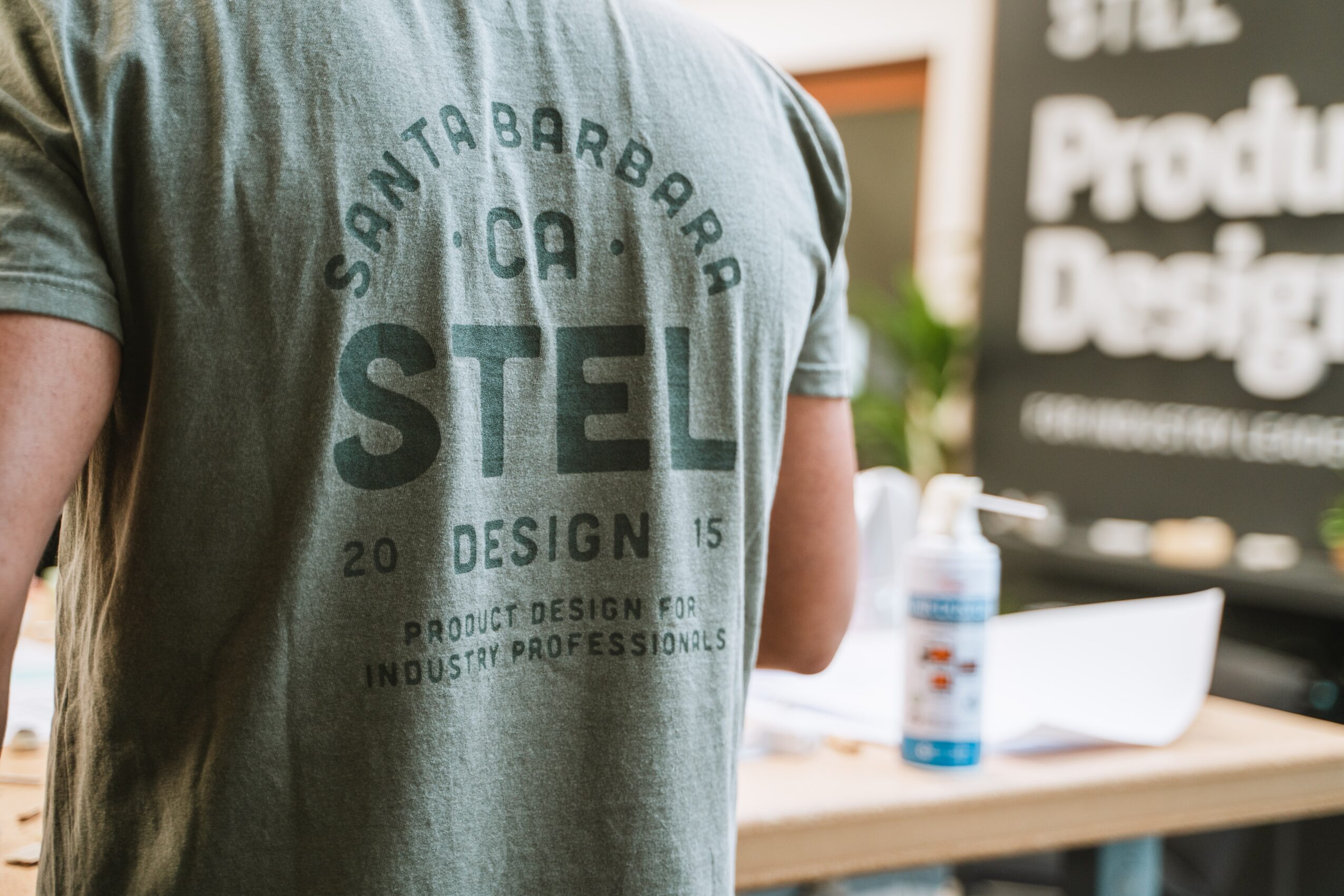 Person wearing a gray T-shirt with text, standing near a table with a water bottle and papers, blurred background.