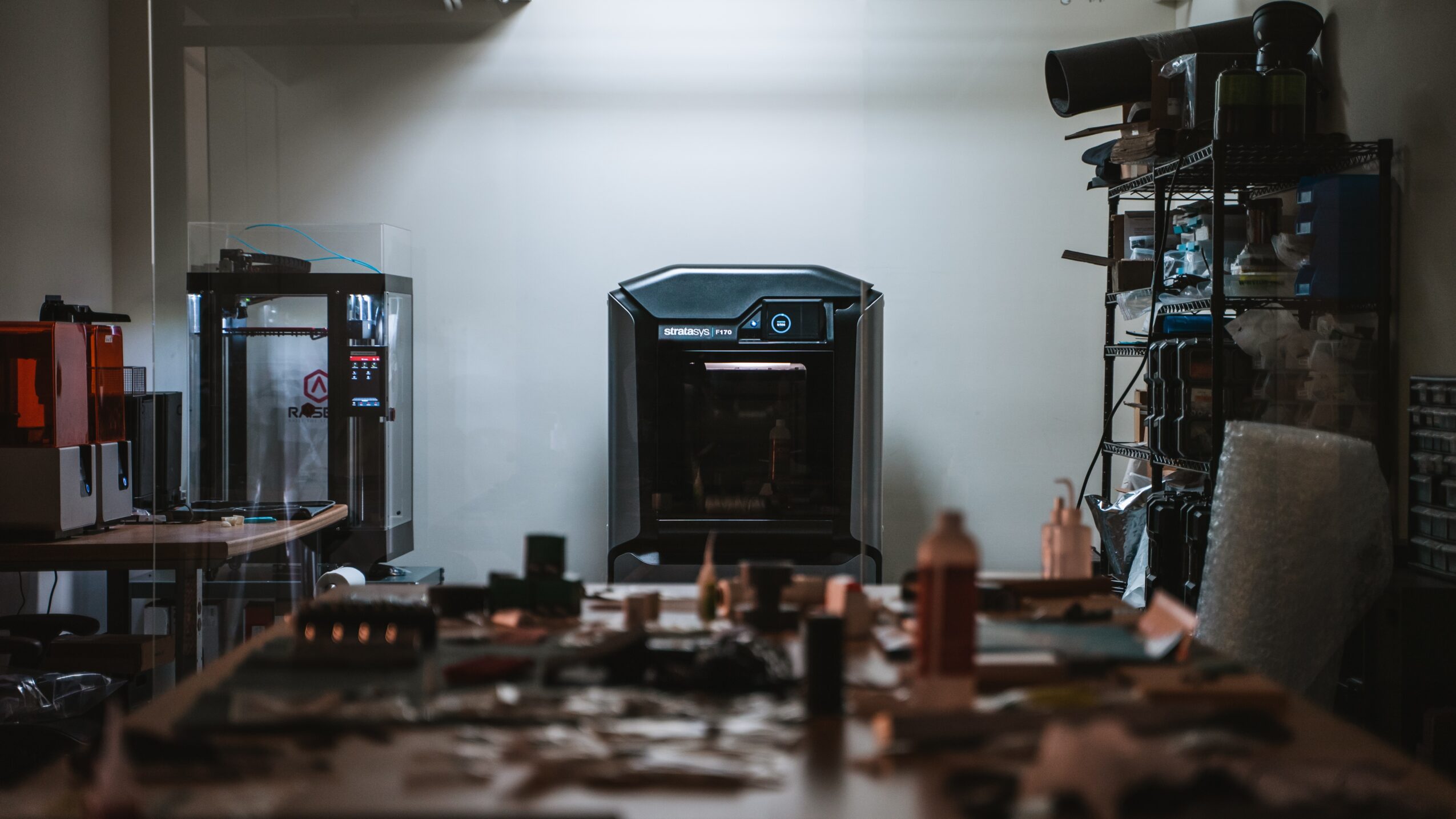 Room with a table, shelves, and a central black appliance, illuminated by overhead lighting, with various objects on the table.