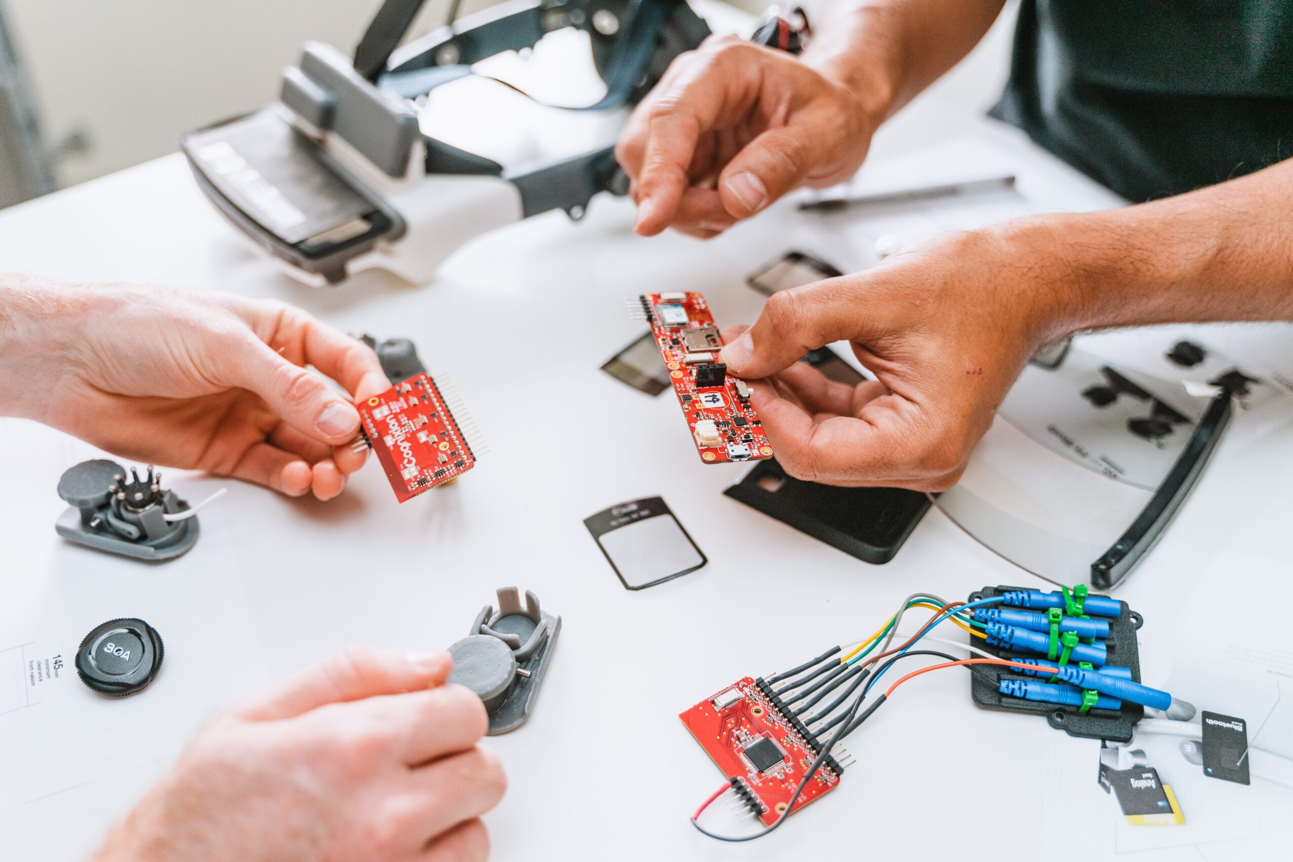 Two people work on electronic components and circuit boards on a white table, with tools and electronic parts visible.