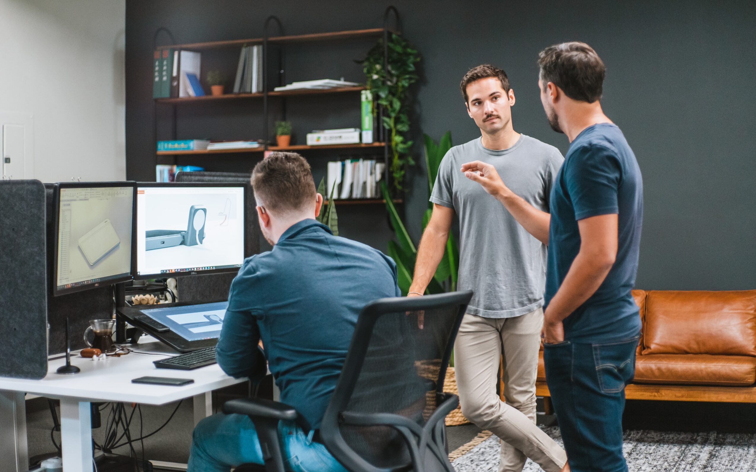 Three men in an office, two standing and one sitting at a desk with computer monitors, discussing work.