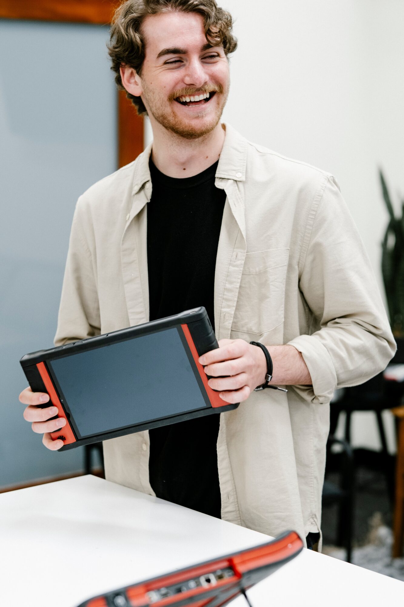 Young man smiling, holding a tablet, wearing a light jacket and black shirt, indoors with a plant in the background.