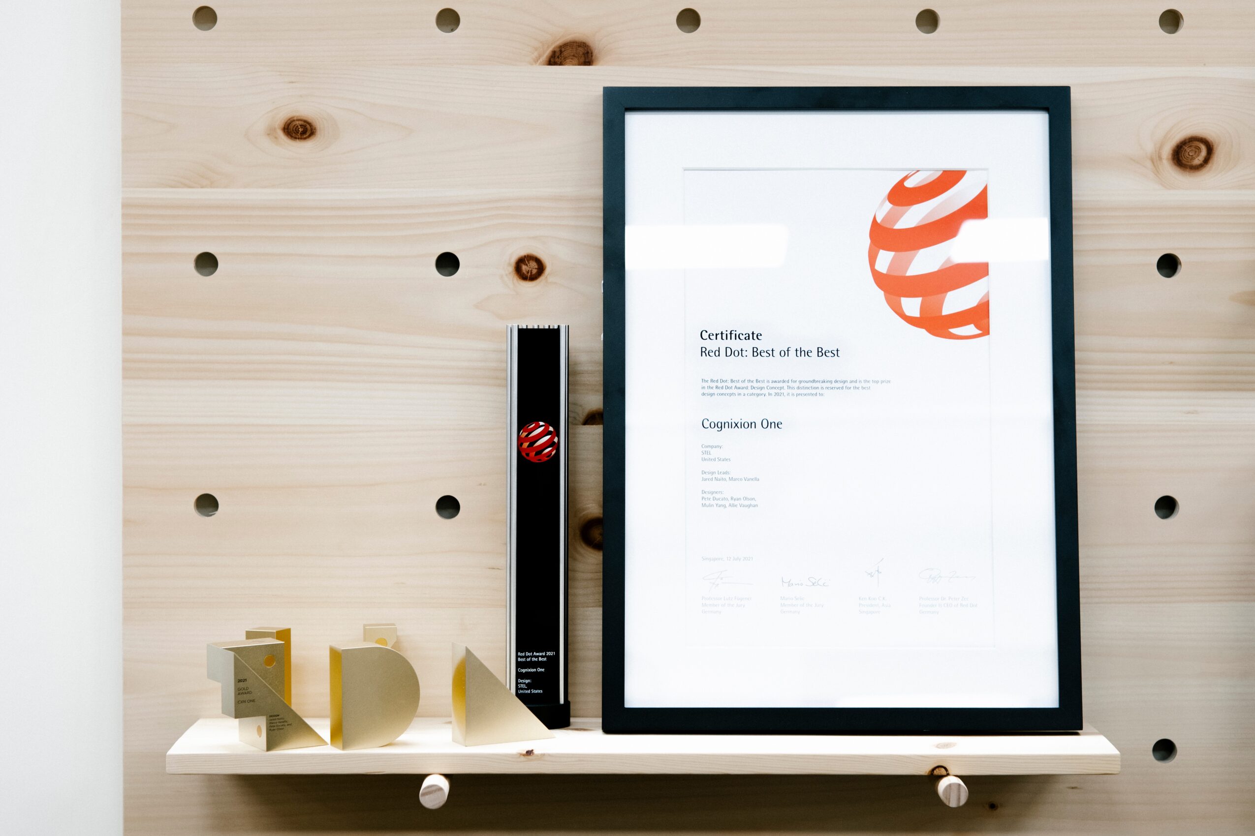 Framed document on a wall with a wooden pegboard background, a black object, and small decorative items on a shelf.