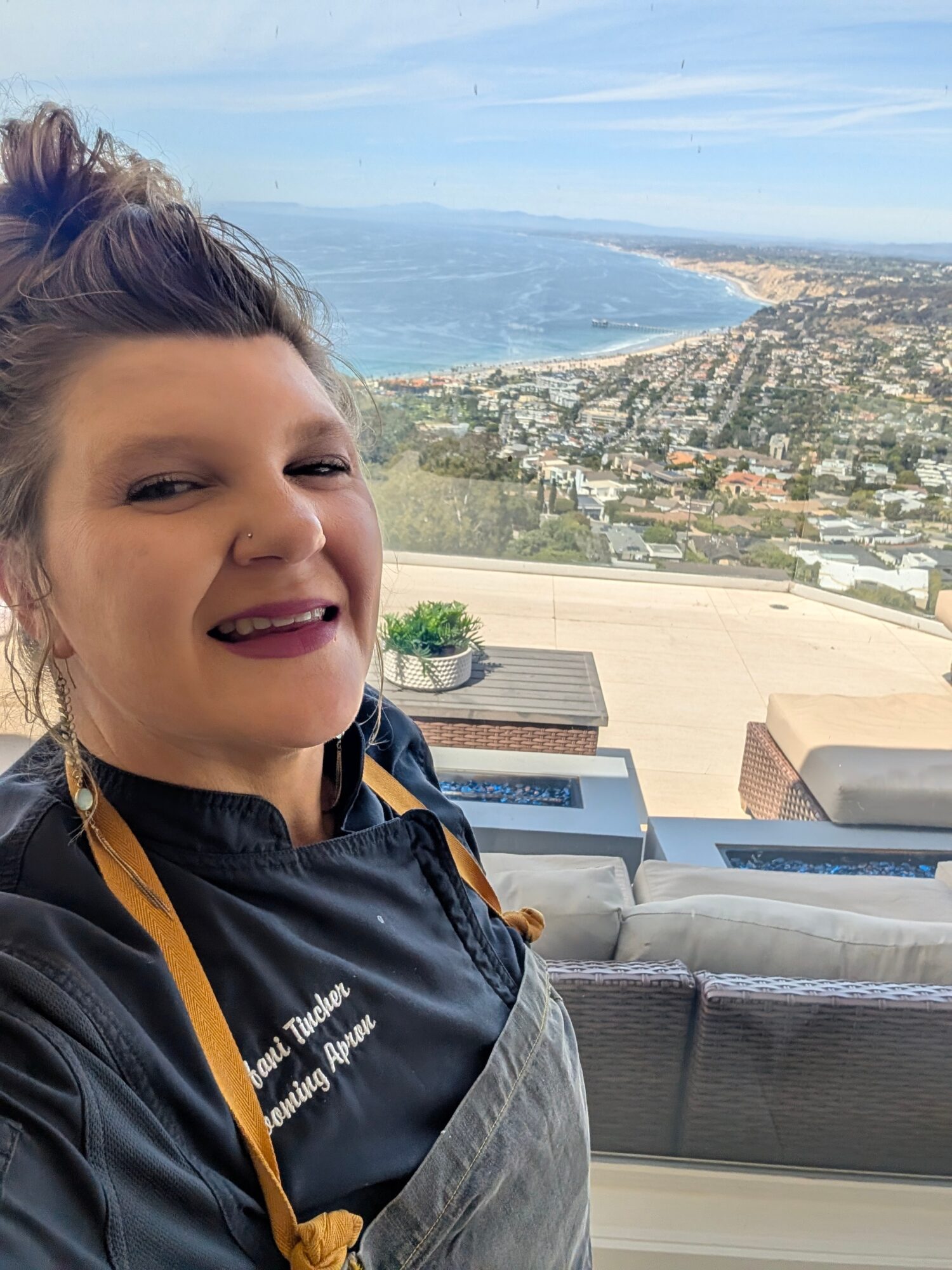 Smiling woman with dark hair in a bun, wearing a black jacket, in front of a large window with a coastal view.
