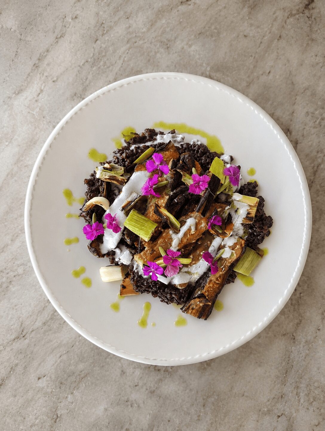Dish with grilled vegetables, black rice, and pink edible flowers on a white plate, on a gray surface.
