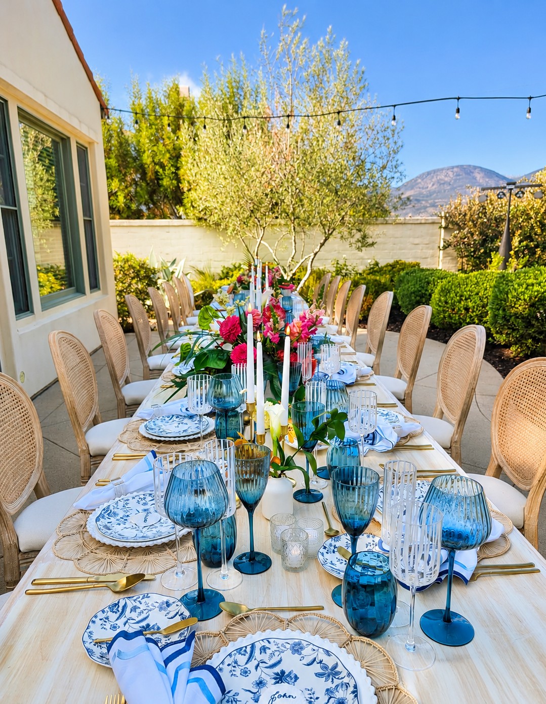 Long outdoor dining table set with plates, glasses, and floral centerpiece, surrounded by chairs, with trees and mountains in background.