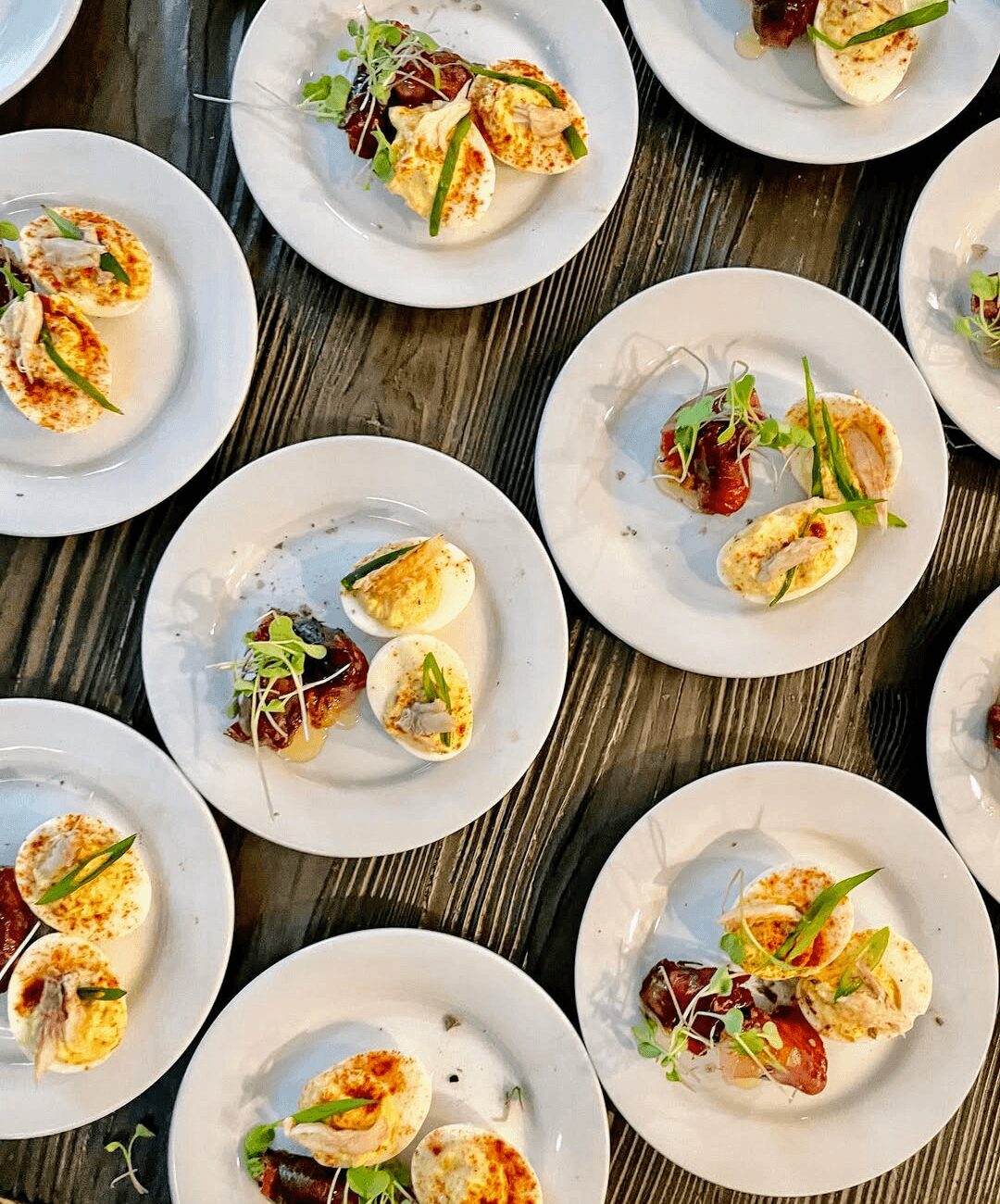 Multiple white plates with appetizers on a dark wooden table, featuring sliced vegetables, herbs, and garnishes.