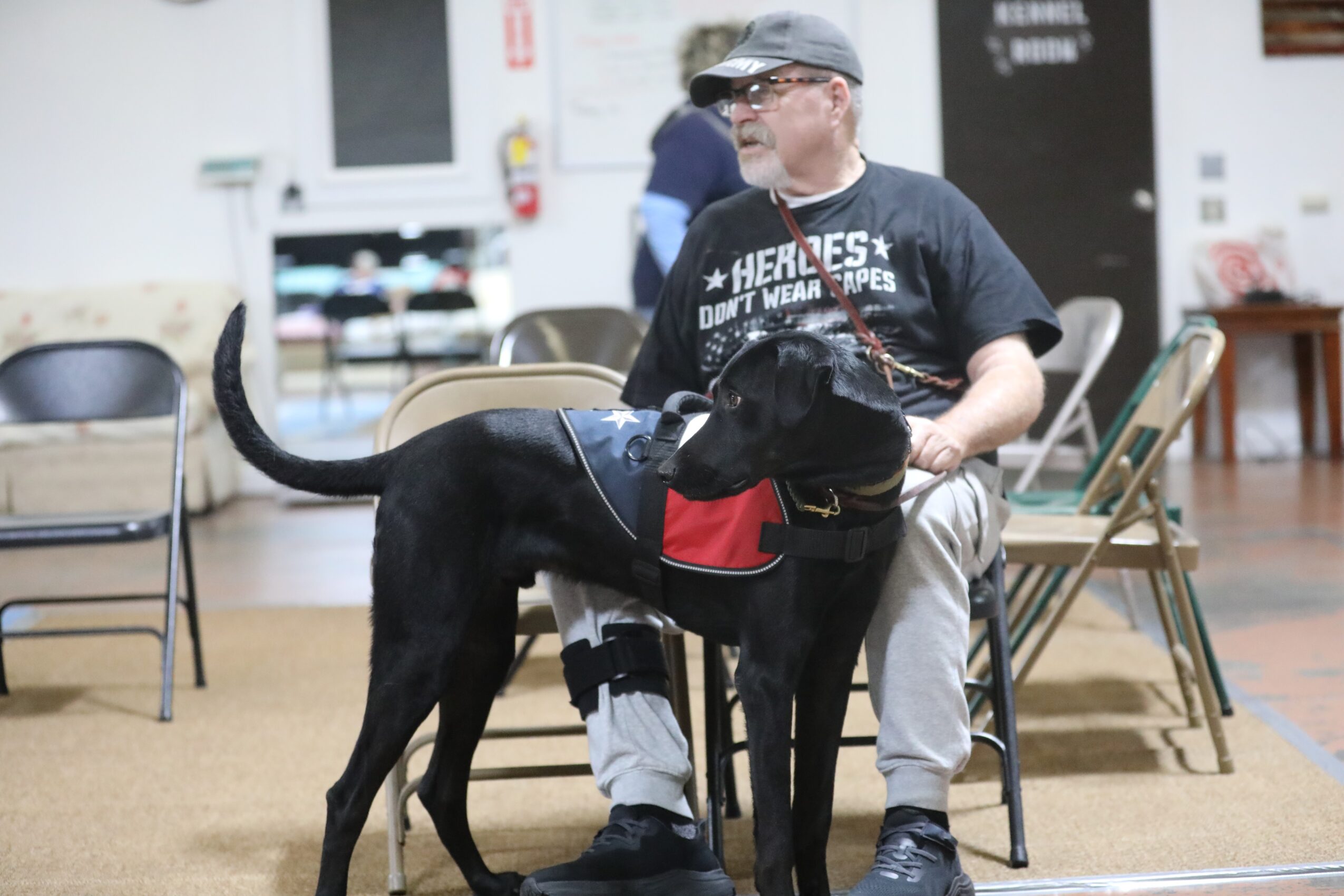 Man sitting on a chair with a black dog wearing a red vest, in an indoor space with chairs and tables.