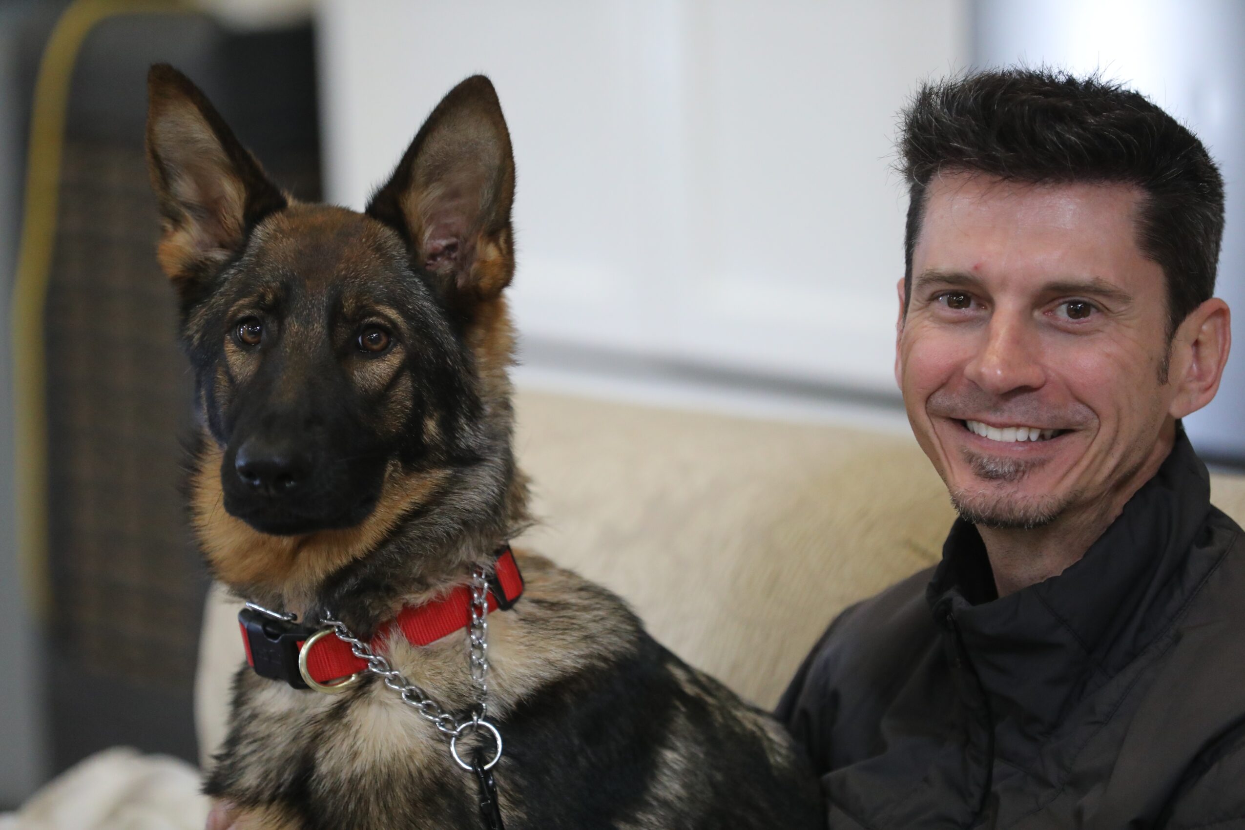 Man smiling next to a German Shepherd dog with a red collar, sitting on a couch indoors.