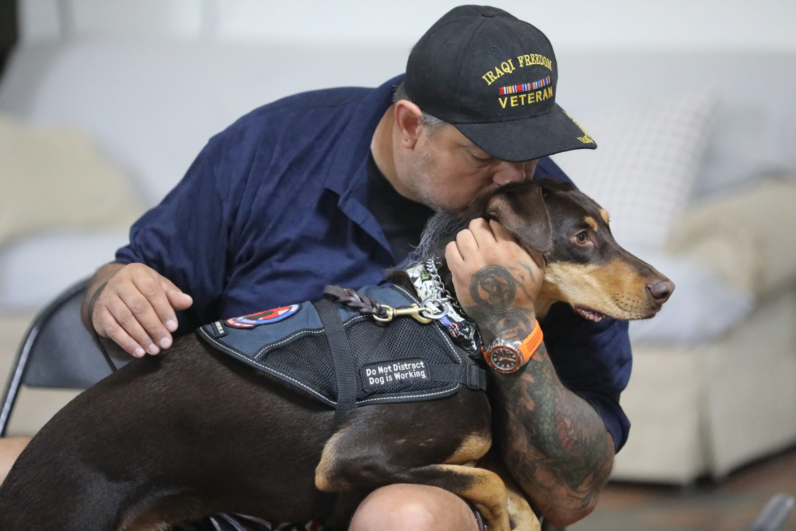 Man in uniform hugging a service dog indoors, both appear close and affectionate.
