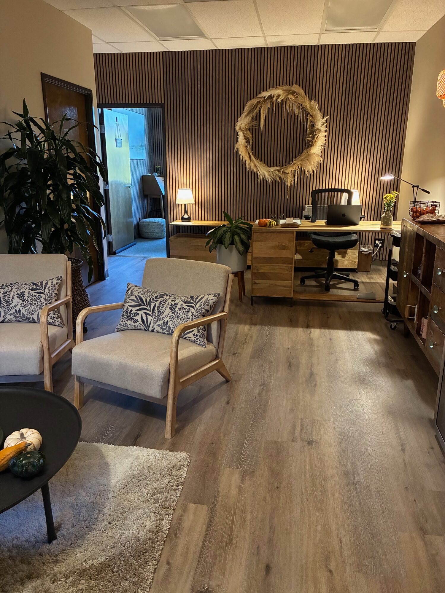 Office or waiting room with chairs, desk, mirror, plants, and a door, decorated in neutral tones.