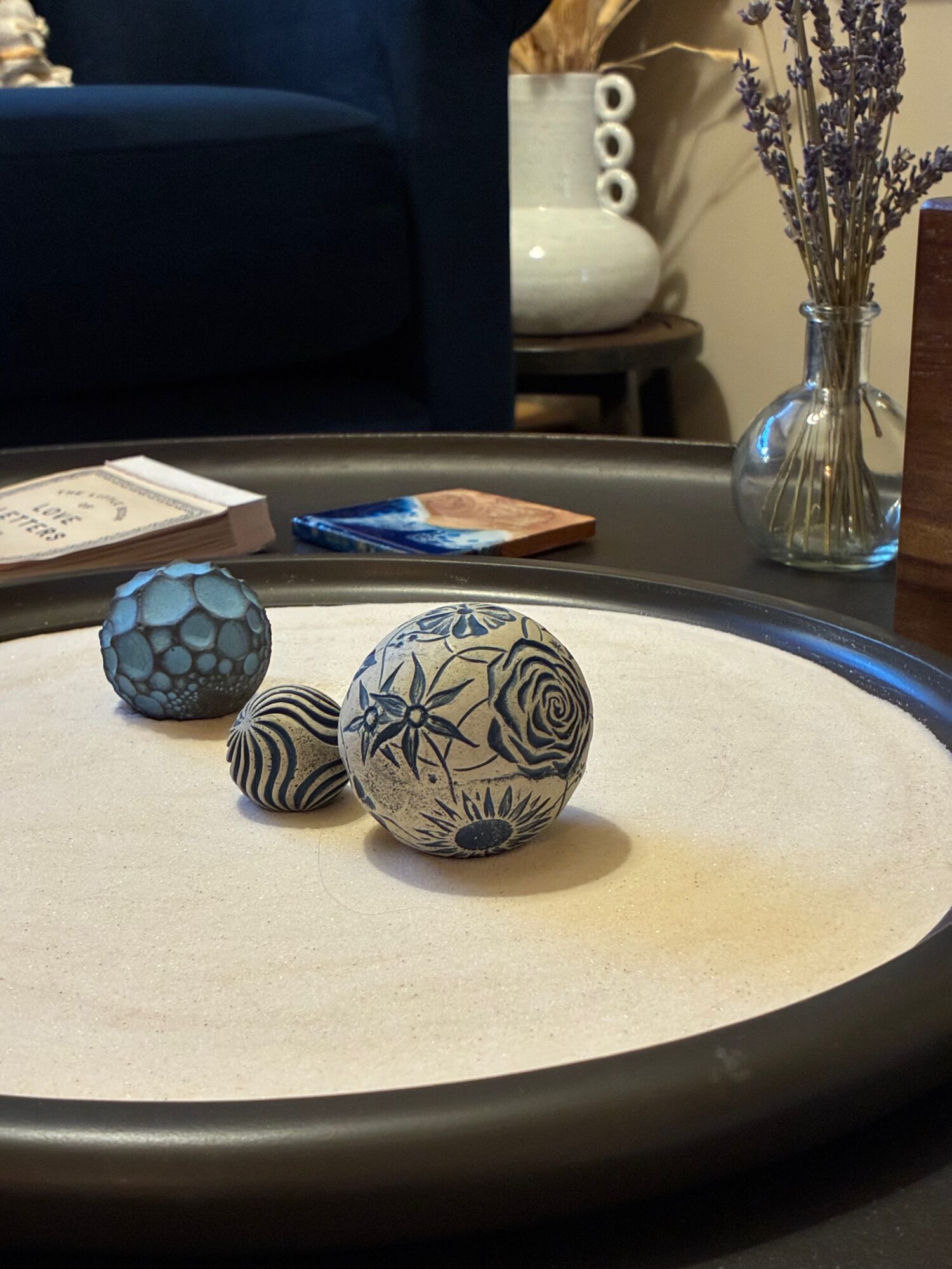 Three decorative spheres on a round table with a white surface, a vase with dried flowers, and a sofa in the background.