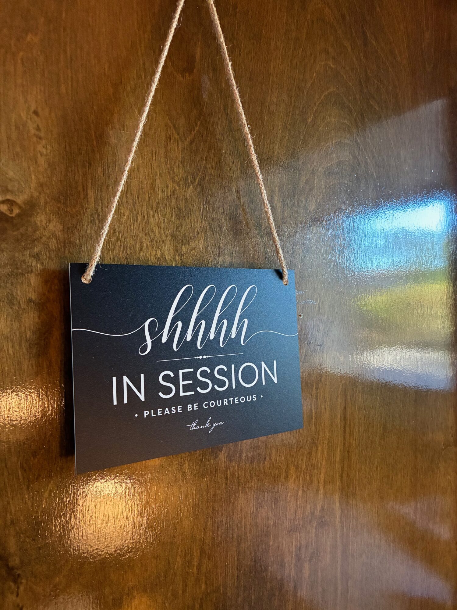 A dark blue sign with white cursive text reading 'shhhh IN SESSION' hangs on a wooden surface with a string.