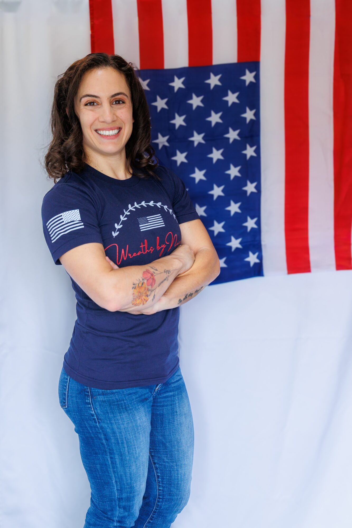 Young woman with curly hair smiling, standing in front of an American flag, arms crossed, wearing a navy T-shirt and jeans.