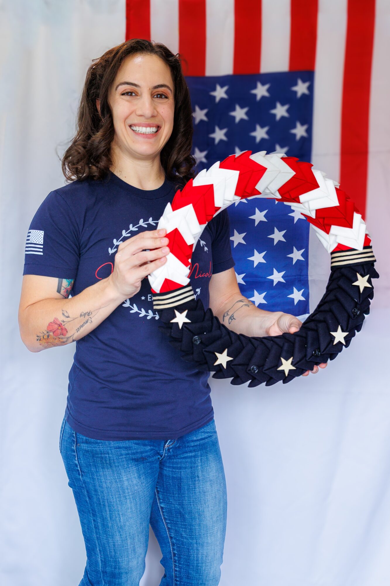 Woman smiling holding a patriotic wreath with stars and stripes, standing in front of an American flag backdrop.