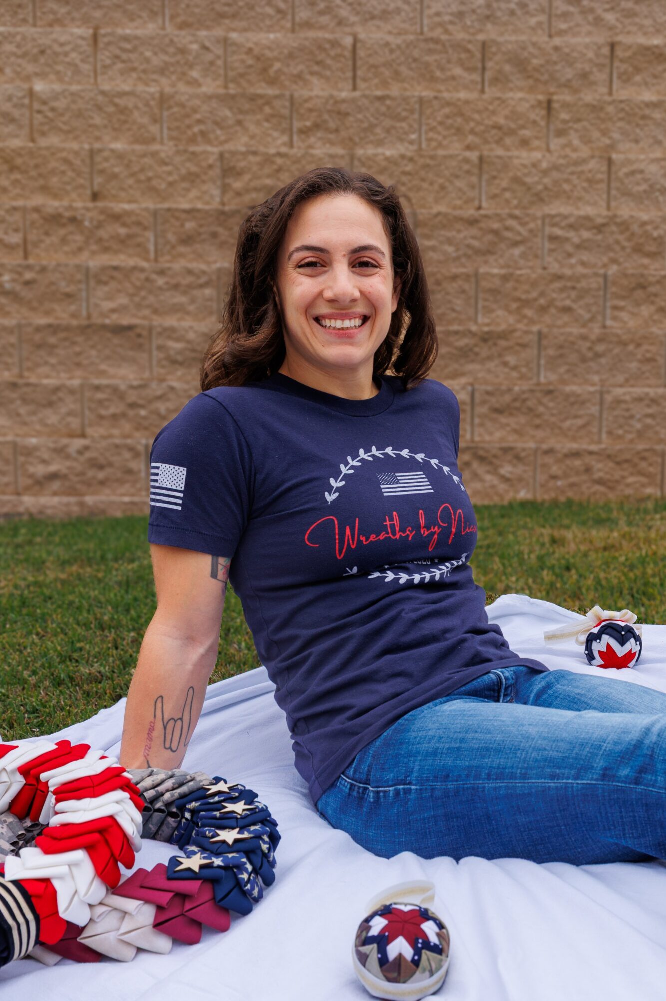 Smiling woman sitting on a bed outdoors, wearing a navy T-shirt and jeans, with a brick wall background.