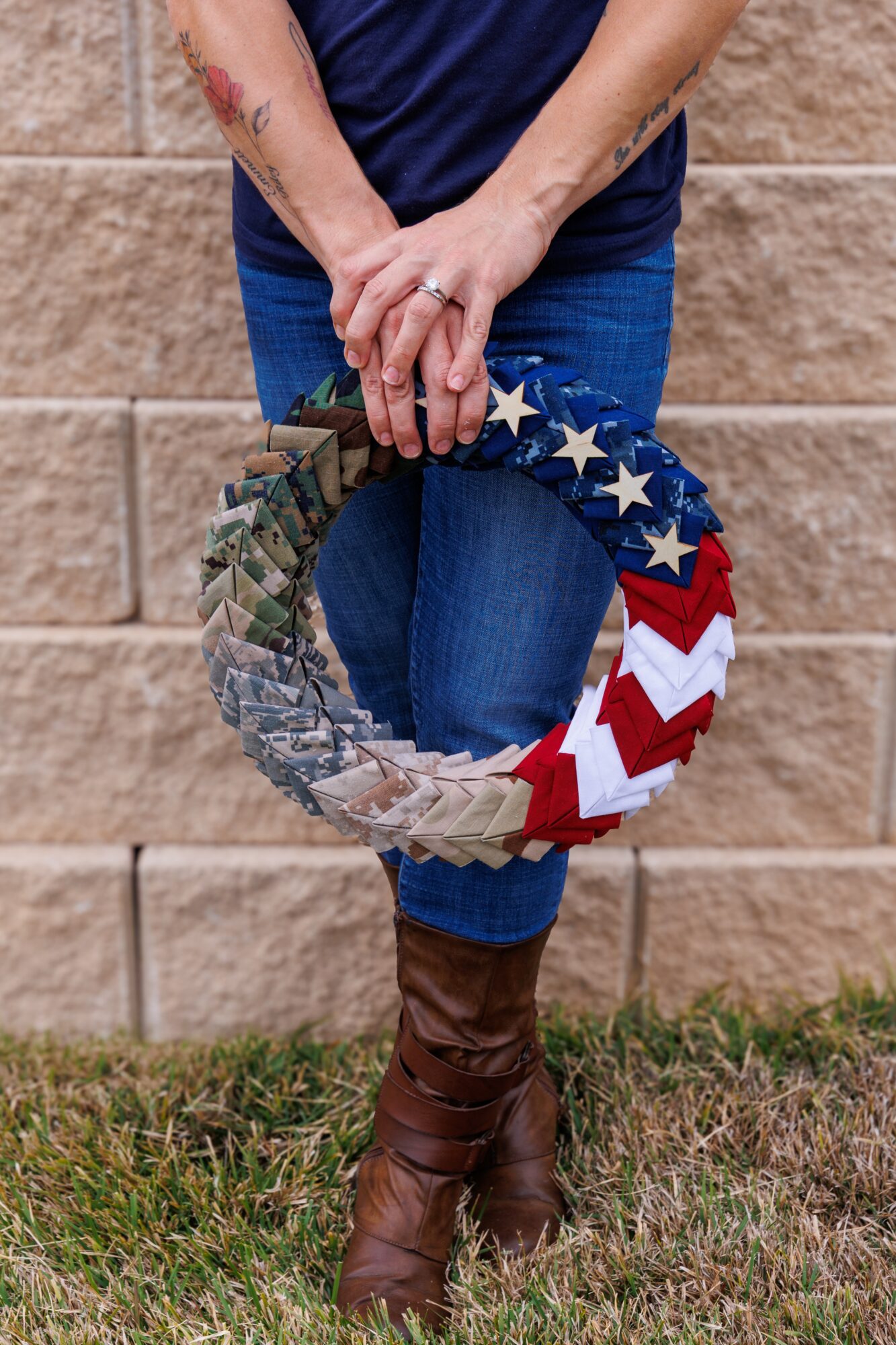 Person holding a patriotic wreath with stars and stripes, standing outdoors against a brick wall, wearing jeans and brown boots.