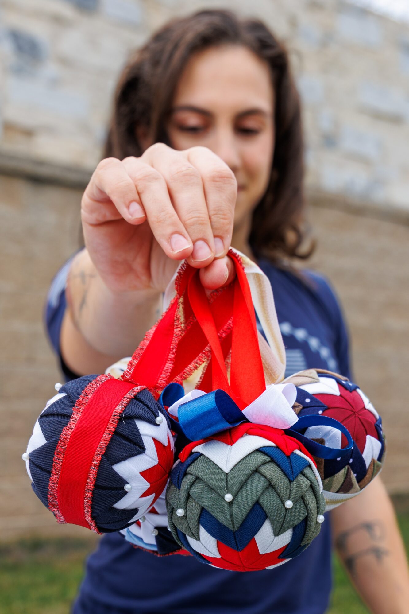 Person holding colorful fabric balls with red ribbons, outdoors with a stone wall background.