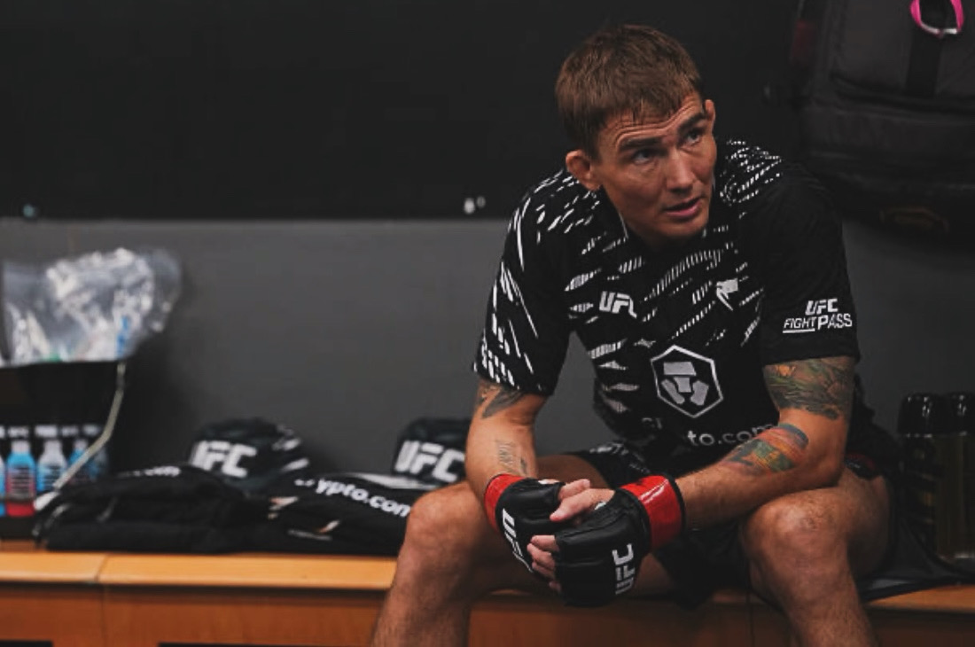 Male mixed martial artist sitting on a bench, wearing gloves and sportswear, in a gym setting.