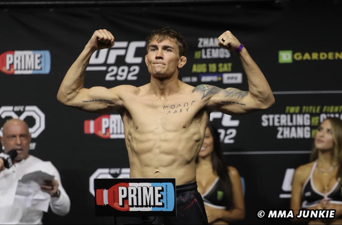 Male fighter with flexed arms on stage, UFC logo behind, people in background, event setting, MMA event, muscular physique.