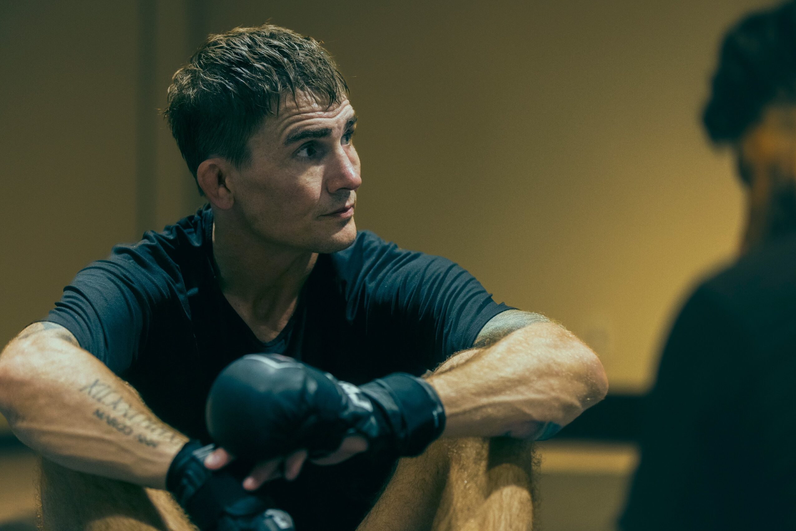 Woman with short hair and tattoos wearing boxing gloves, sitting and talking to a person with curly hair, in a room with yellowish lighting.