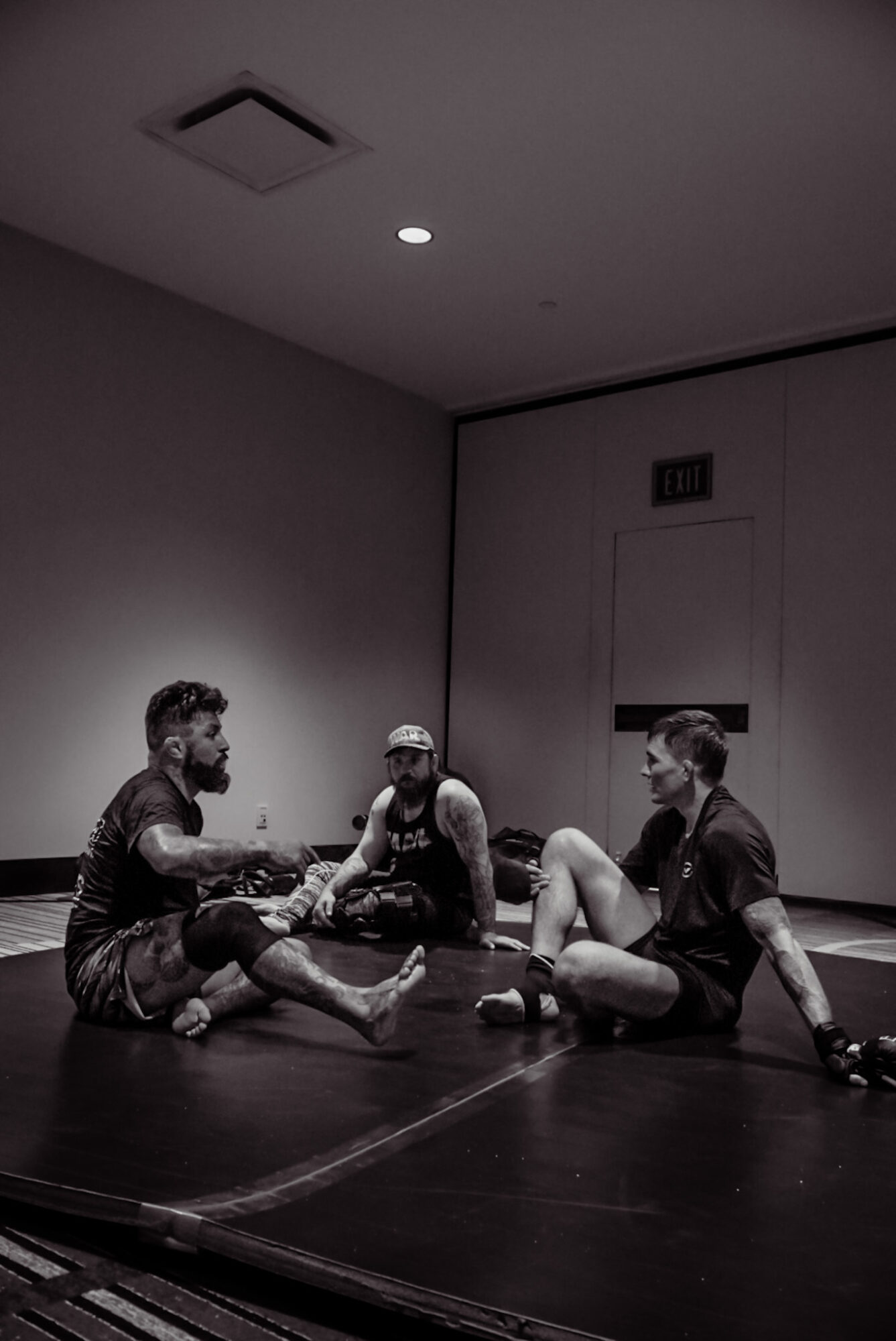 Three people sitting on a stage floor in a room with plain walls, engaged in conversation or activity.