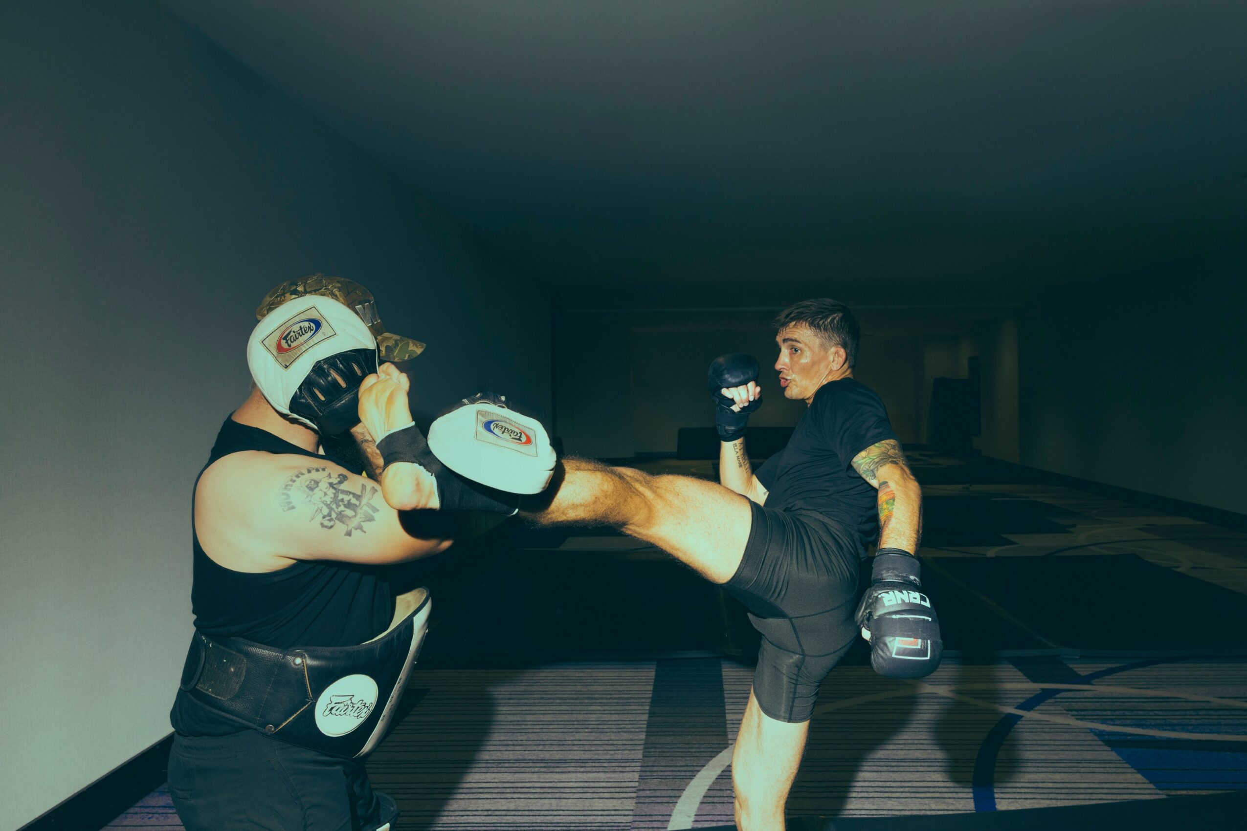 Two women in boxing gear sparring indoors, one delivering a kick to the other's midsection, both wearing gloves and protective headgear.