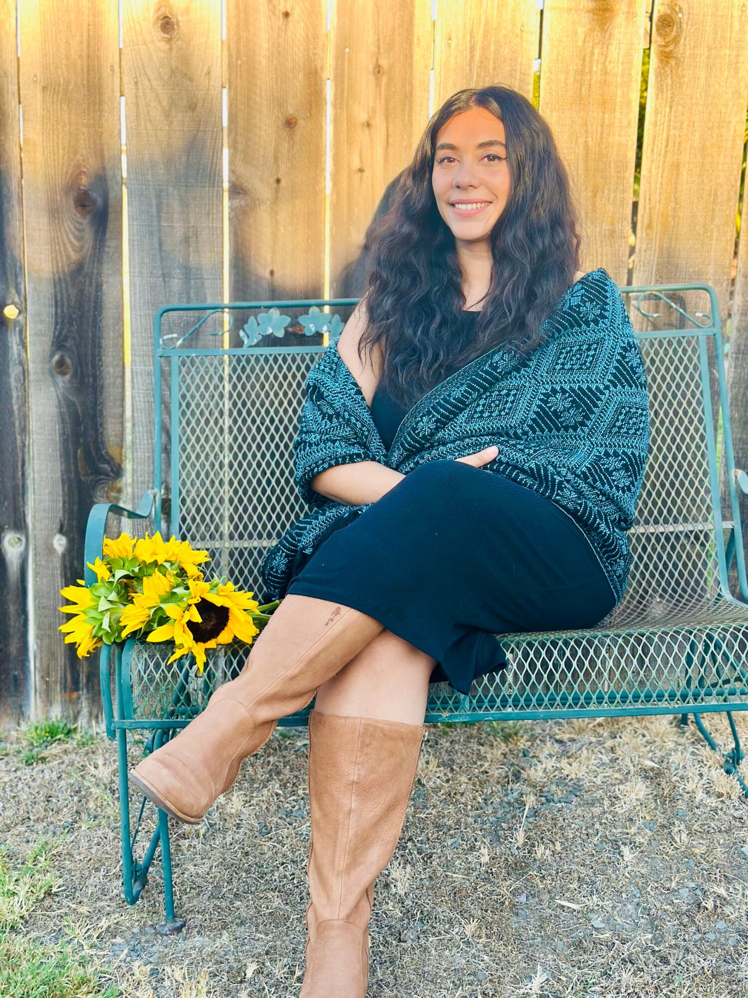 Young woman with long curly hair sitting on a metal bench outdoors, smiling, with a wooden fence in the background and a basket of sunflowers nearby.
