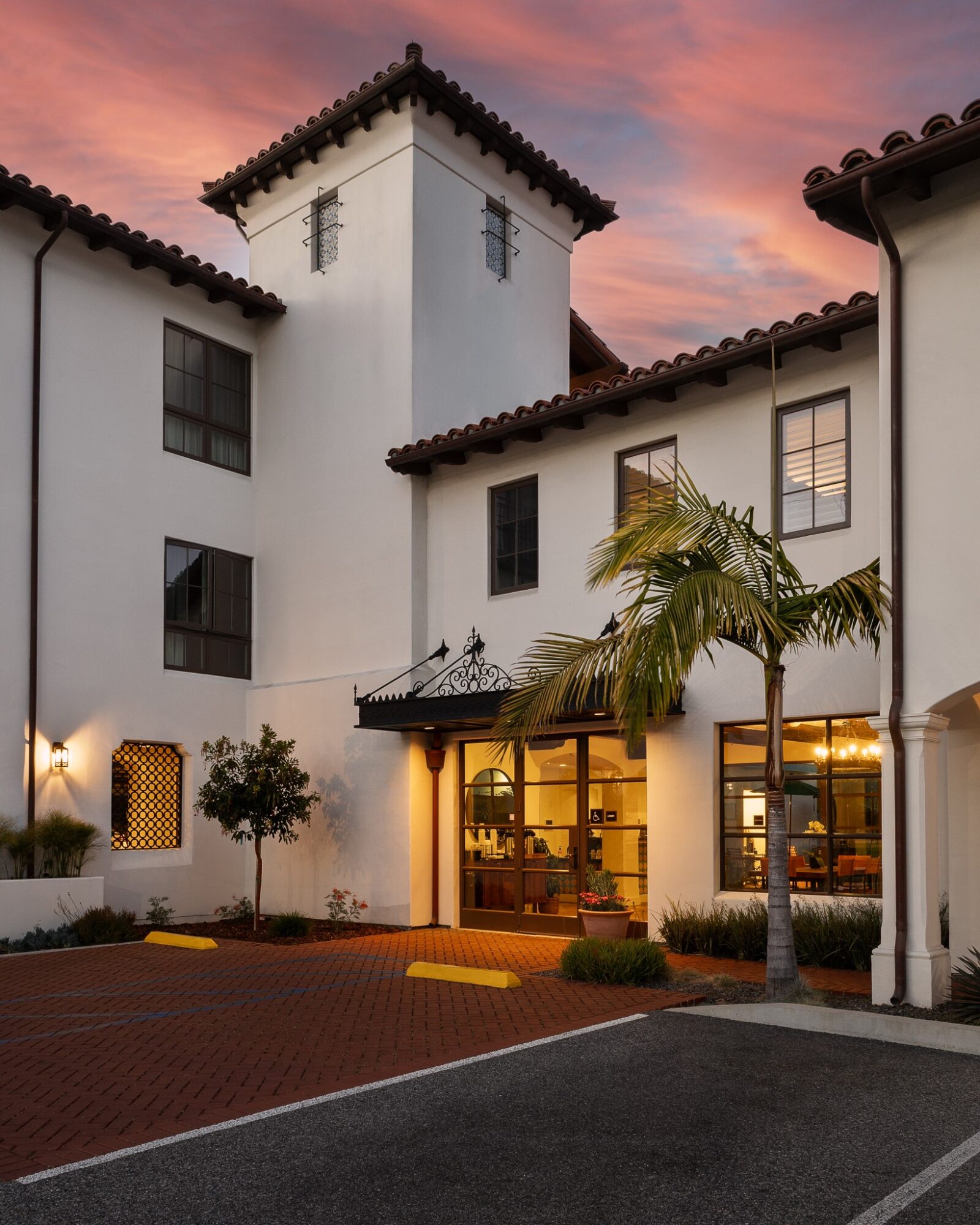 White building with a tower, palm tree, and sunset sky, with a brick parking area in front.