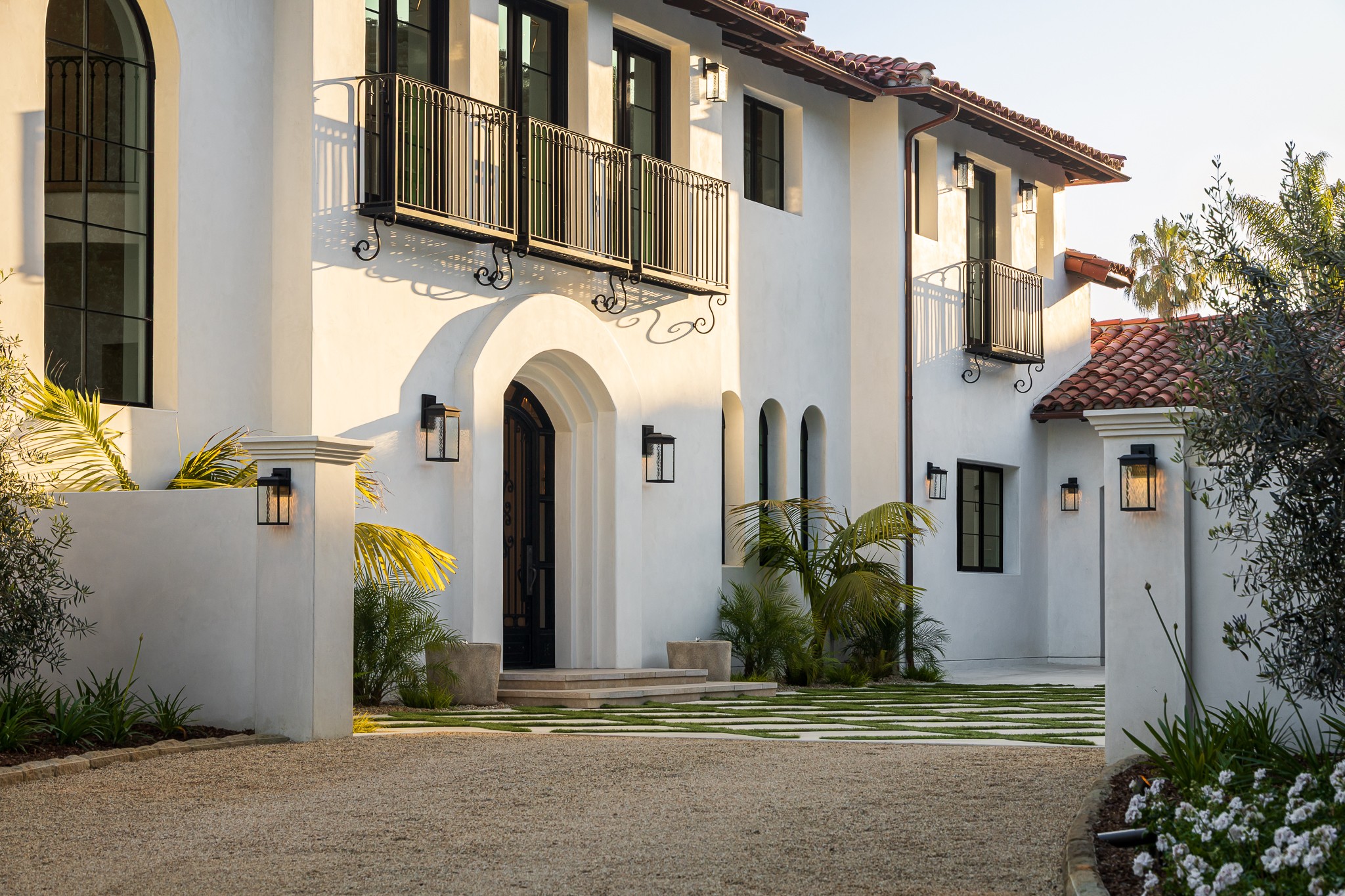 White two-story house with balconies, arched doorway, and outdoor lighting, surrounded by greenery and a driveway.