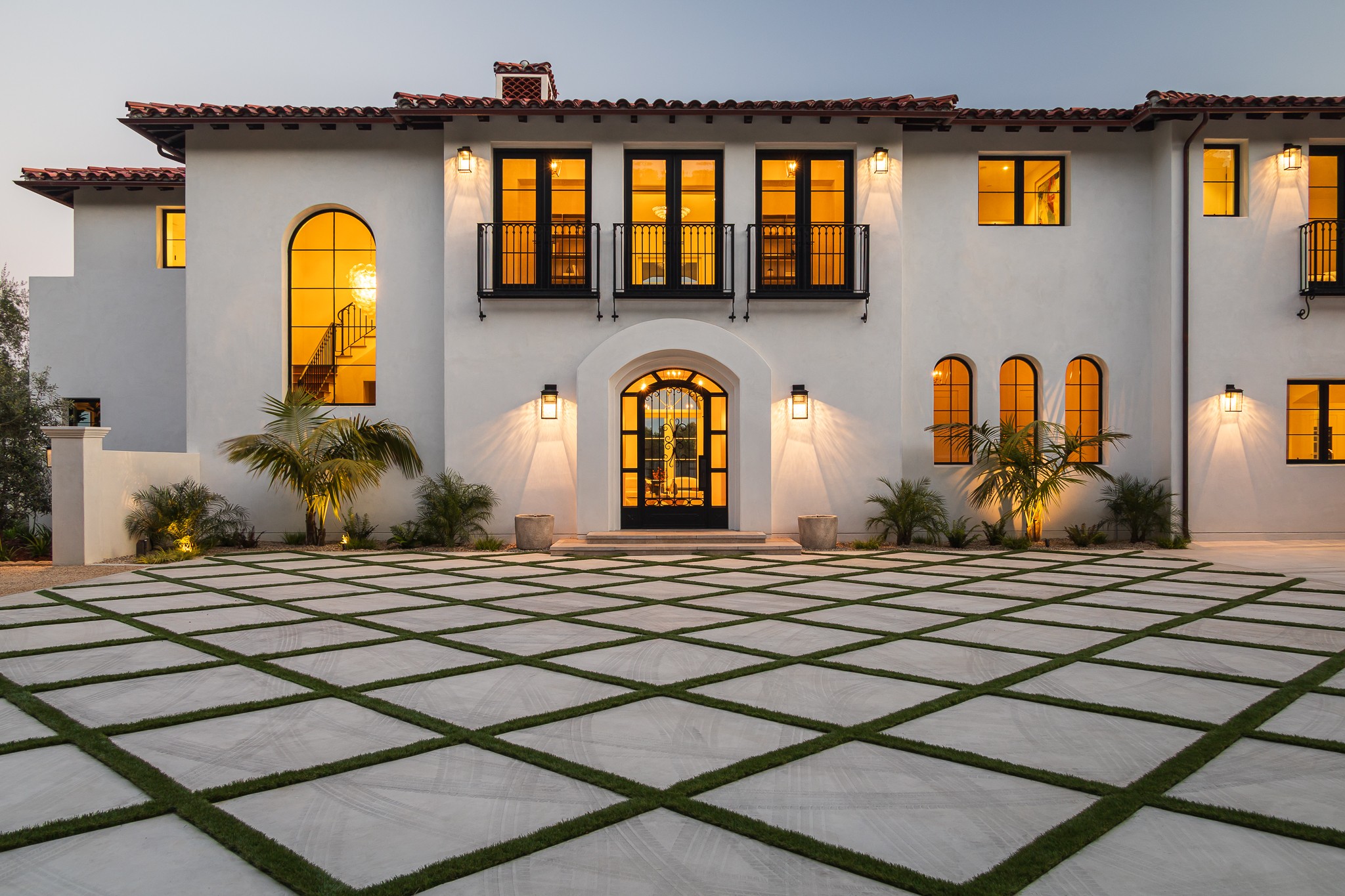 Large white house with arched and rectangular windows, black balcony, and a central door, surrounded by a patterned driveway and palm trees.