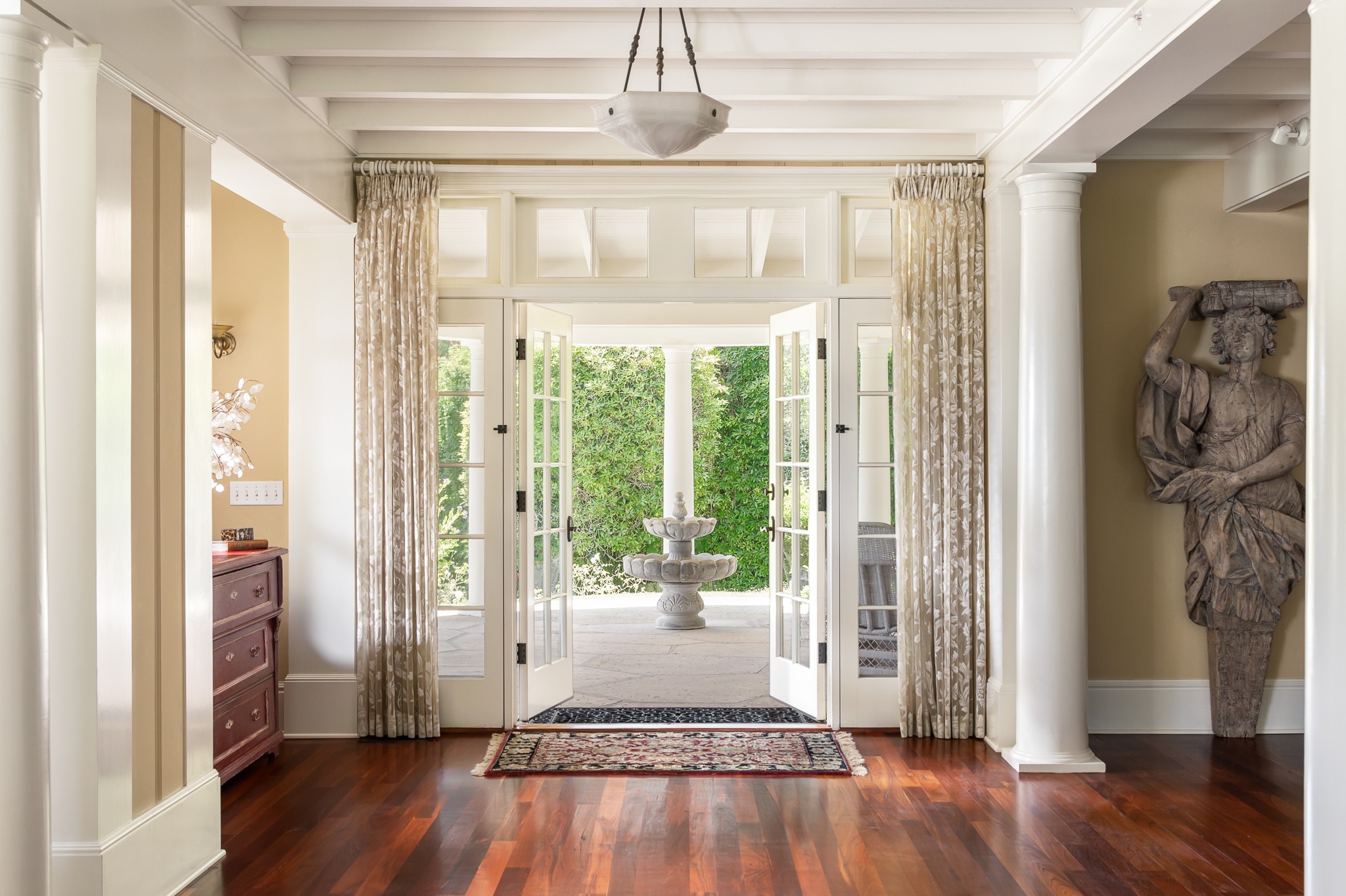 Open double doors leading to a garden with a fountain, framed by curtains and columns, with hardwood floors and a rug.