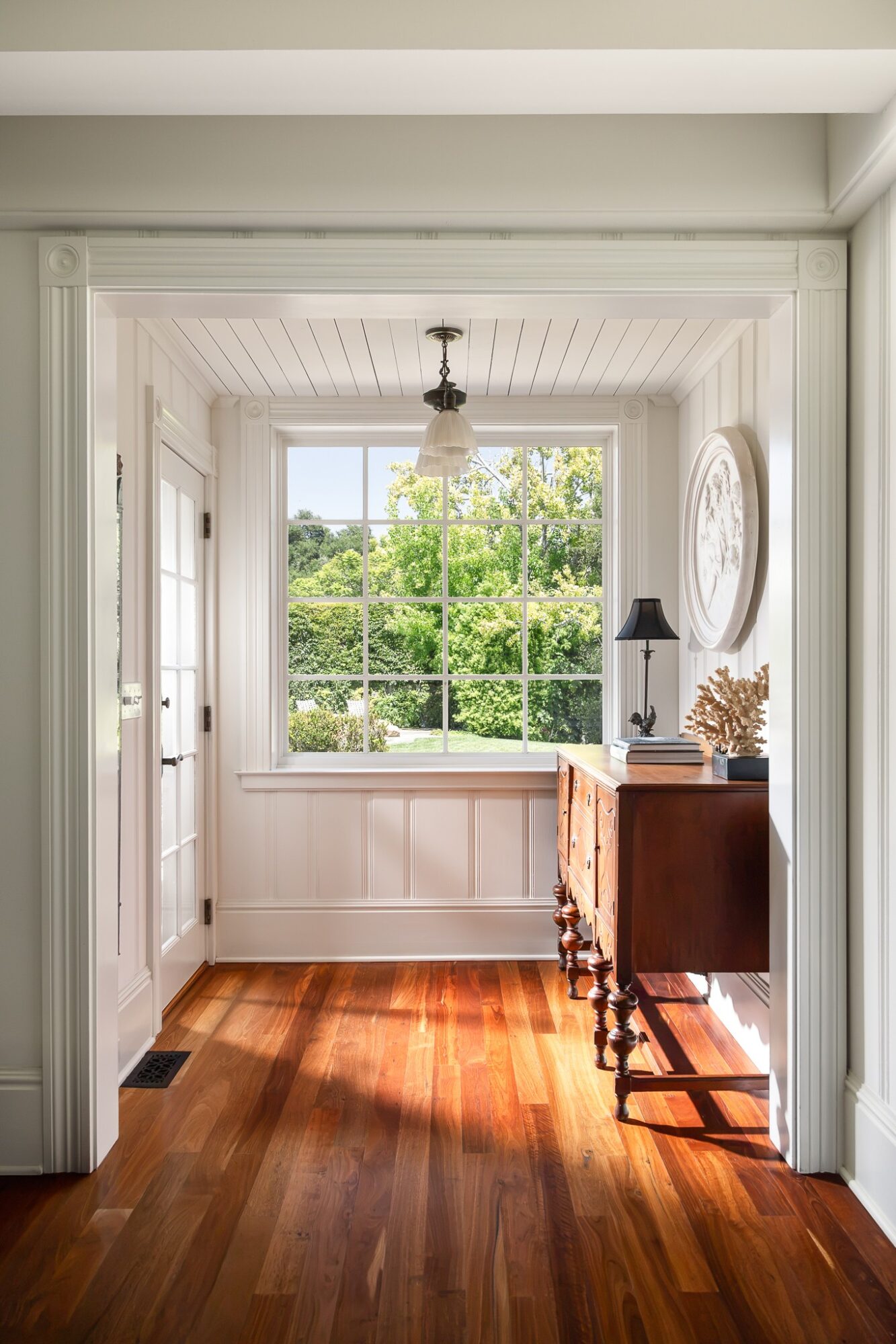 Sunlit room with large window, wooden floor, and a small wooden table with a lamp and decorative items.