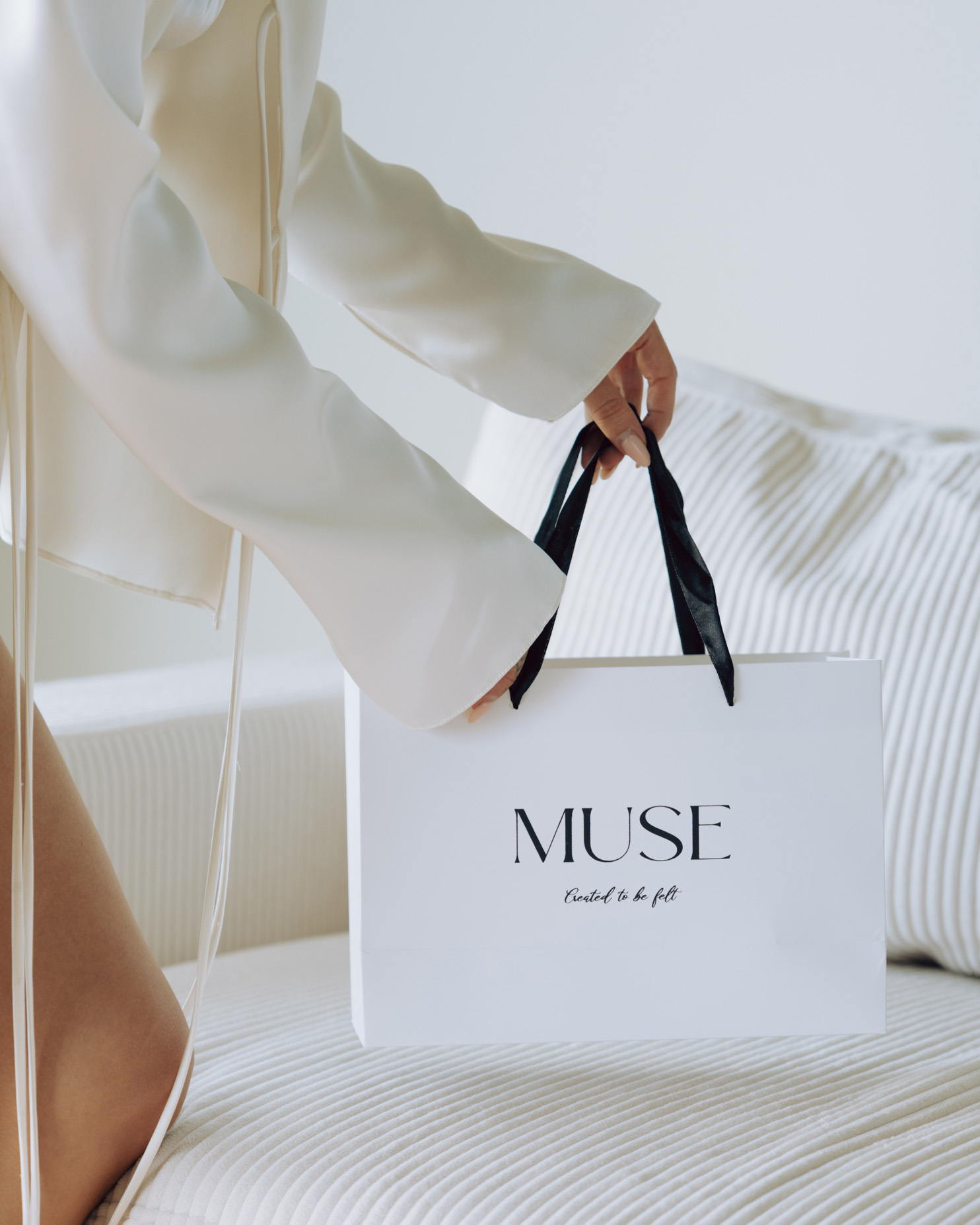 Person holding a white shopping bag with black handles on a bed with striped bedding.