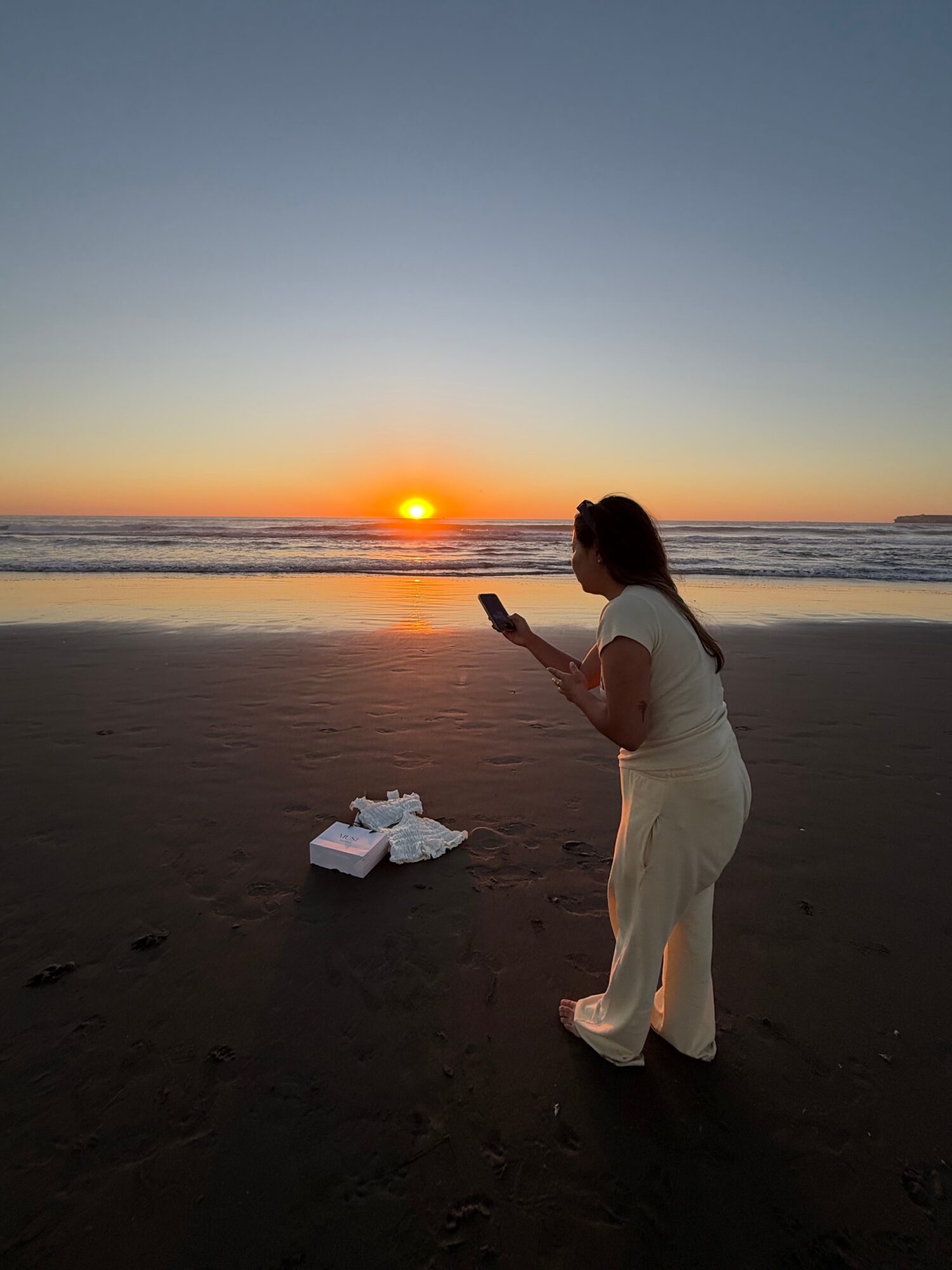 Person in white clothes standing on beach at sunset, looking at phone, with food containers on sand nearby.