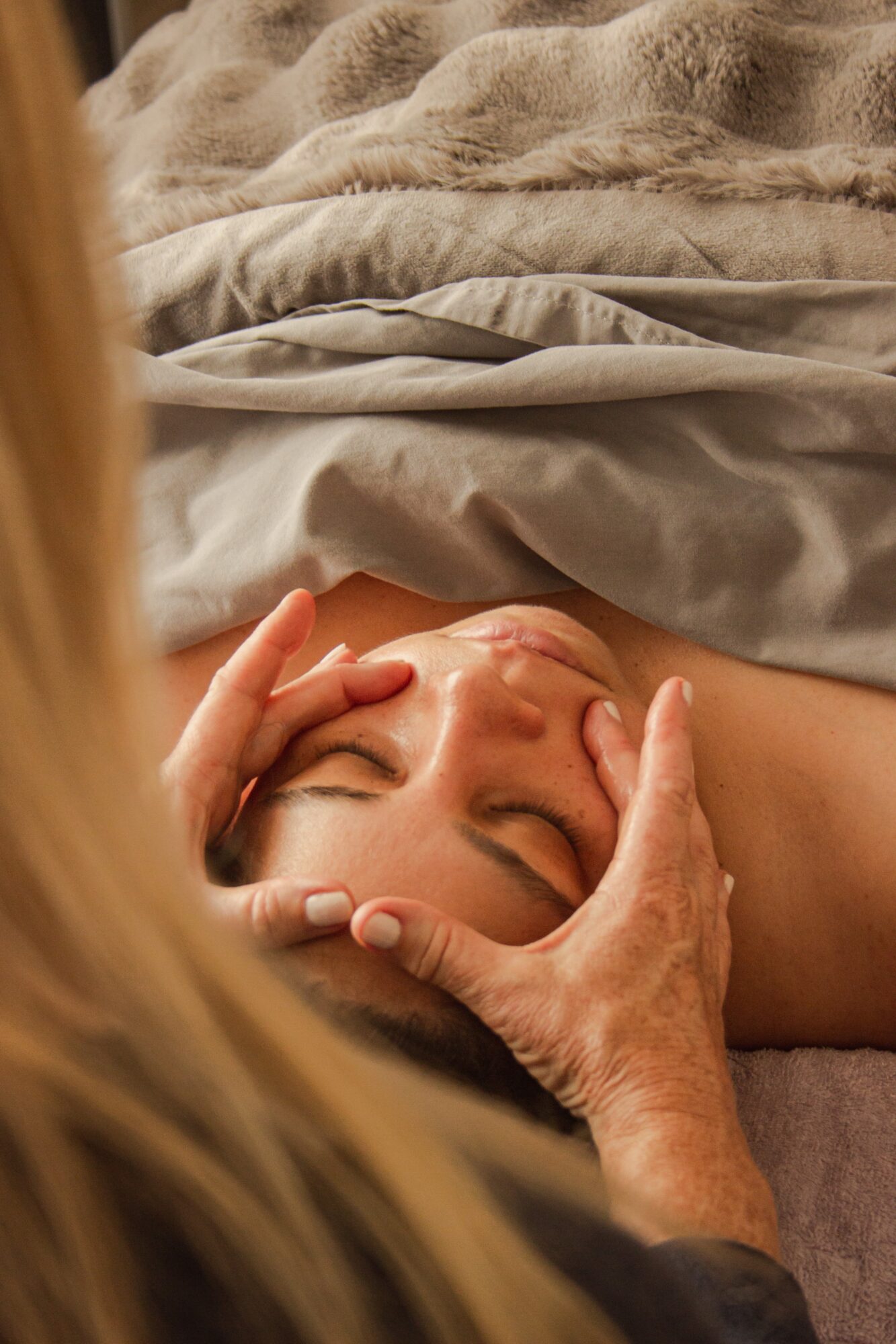 Person lying in bed with hands gently touching their face, eyes closed, surrounded by soft blankets.