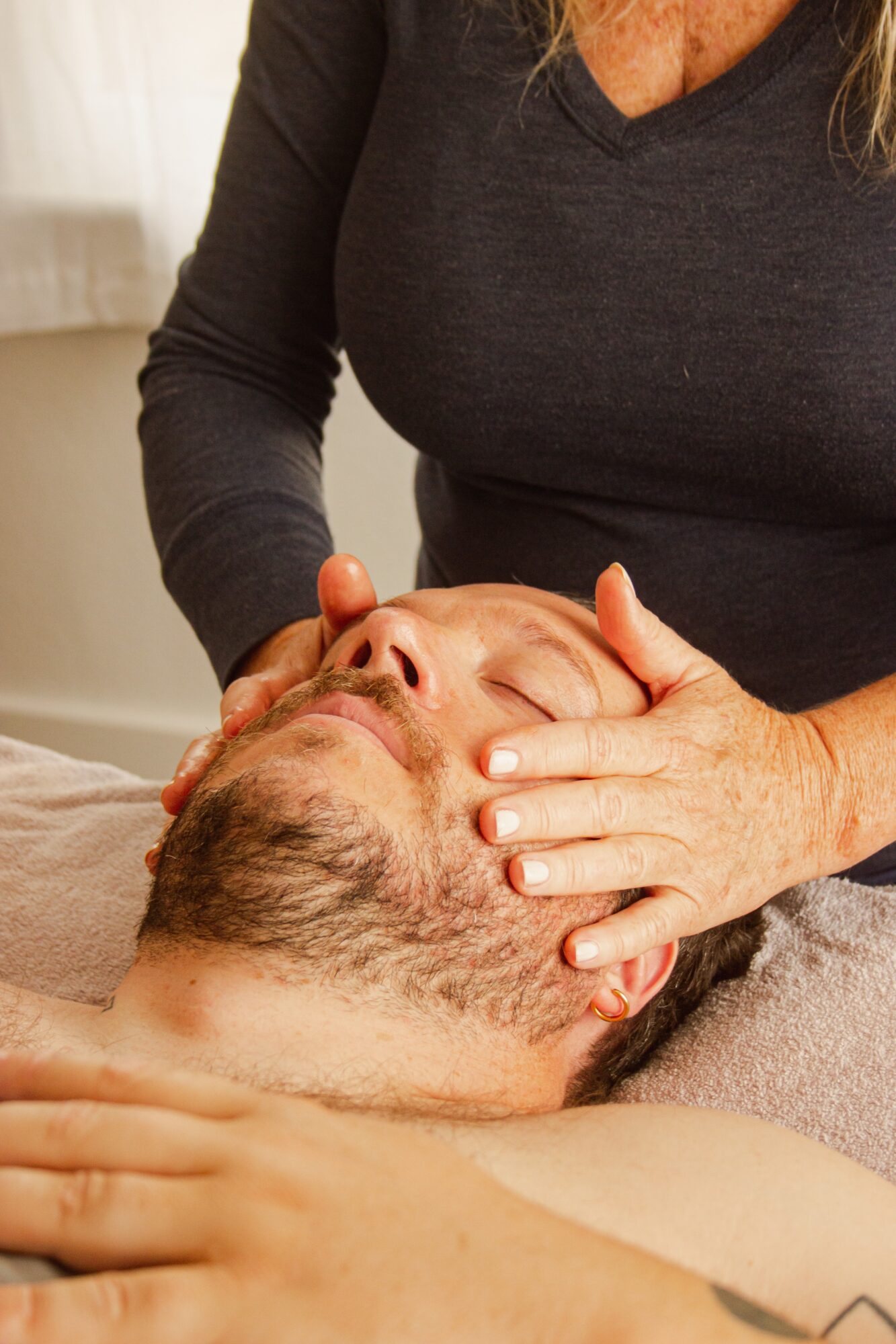 Person receiving facial massage from a therapist, lying down with eyes closed, in a room with natural light.