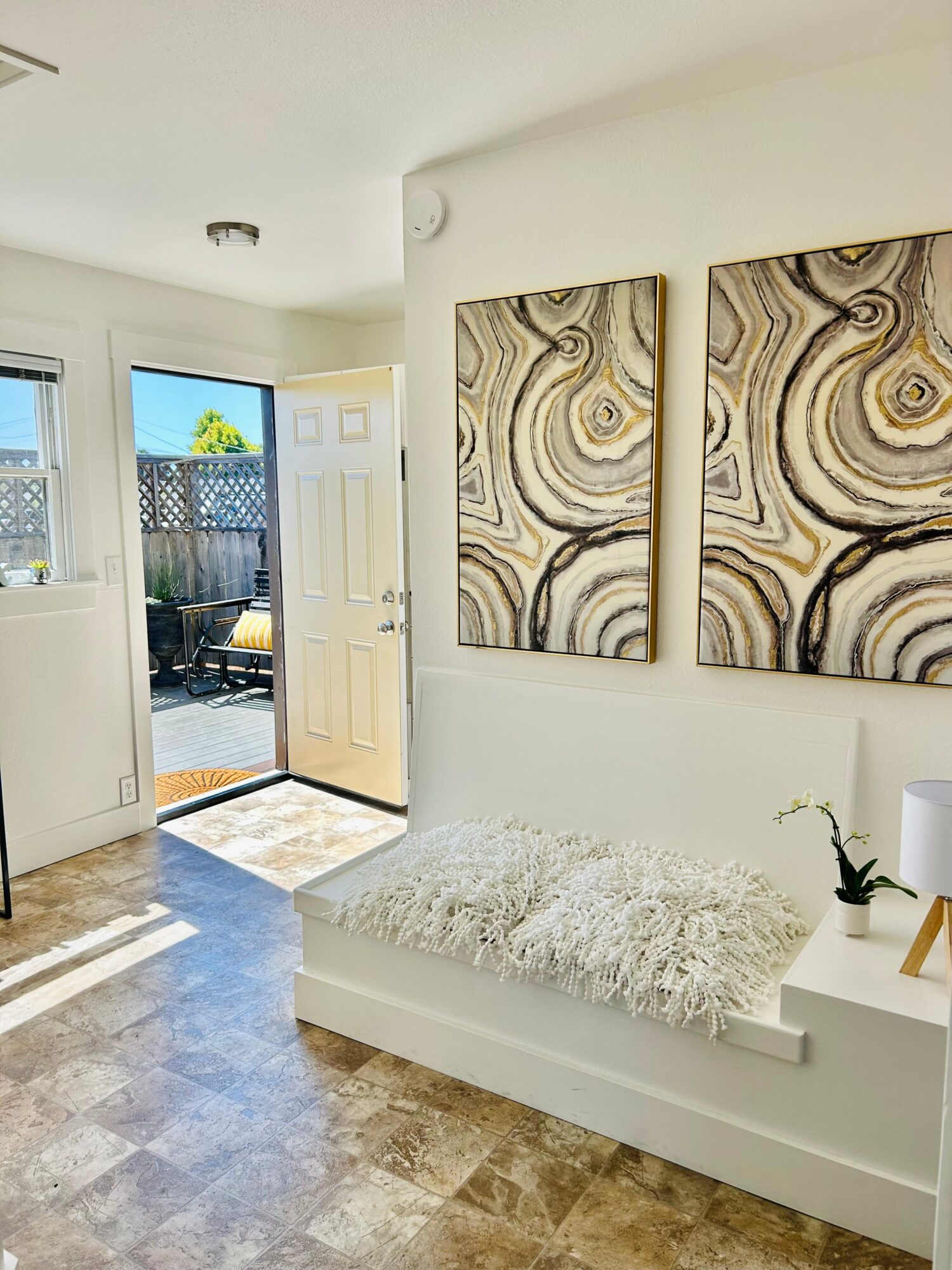 Sunlit entryway with open door, two abstract wall art pieces, a white bench with a fluffy cushion, and a small plant.