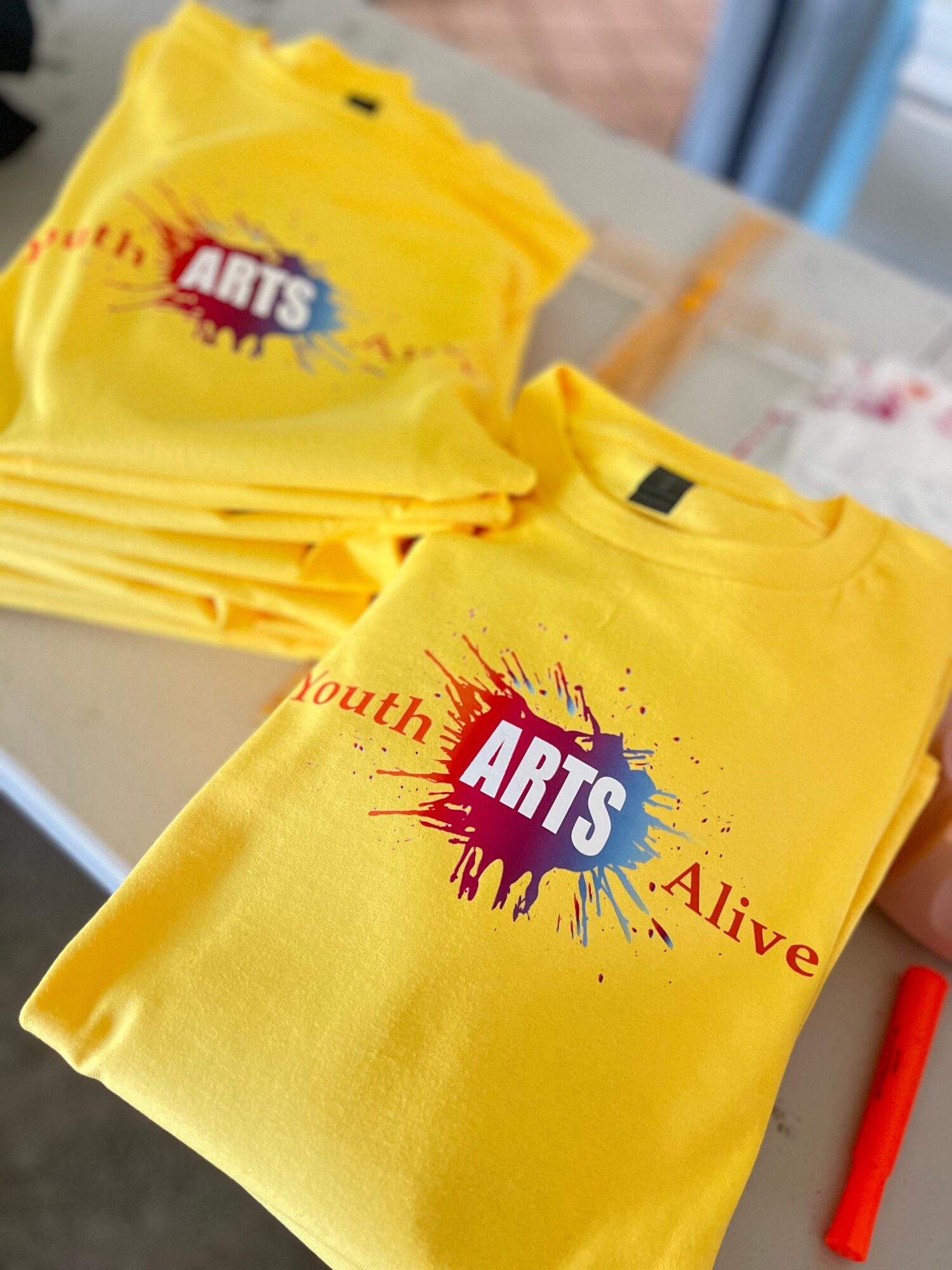 Two yellow T-shirts with a colorful logo reading 'ARTS' and text 'Youth Alive' on a table, with a red marker nearby.