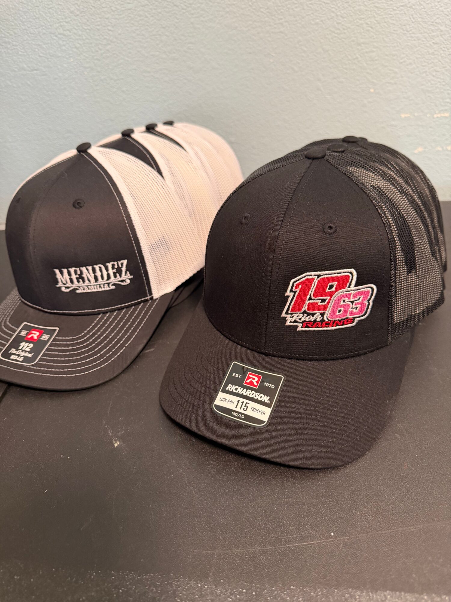 Two baseball caps, one black with a logo and the other white with a logo, placed on a dark surface against a light wall.